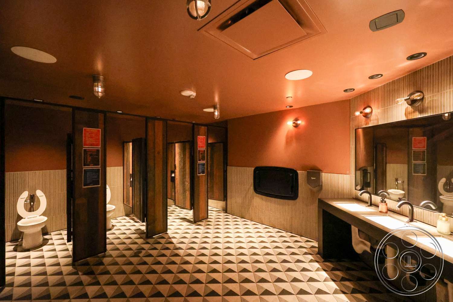 Club 74 - textured plaster walls, bright ceiling lights, interior restroom space, triangular patterned tile flooring, wooden stall partitions, modern sink fixtures, dual-flush toilets, ambient wall lighting