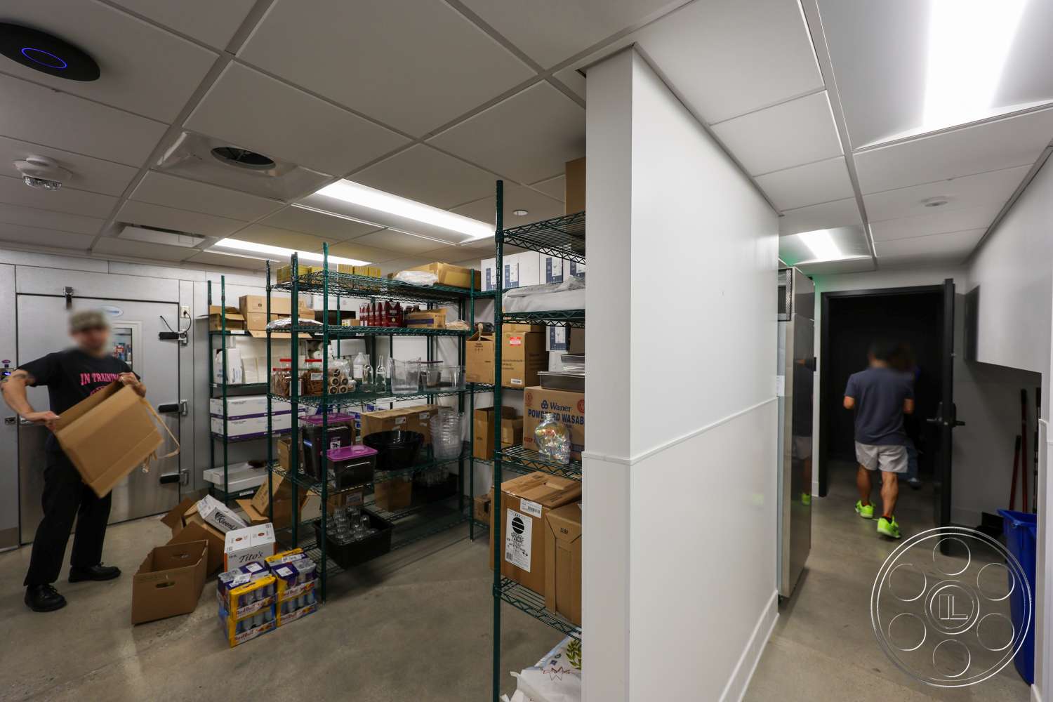 Club 74 - concrete flooring, metal shelving units, glassware storage, cardboard boxes, dimly lit corridor, white painted walls, drop ceiling with fluorescent lighting, organized storage space, storage room, commercial kitchen supplies, refrigerated storage unit