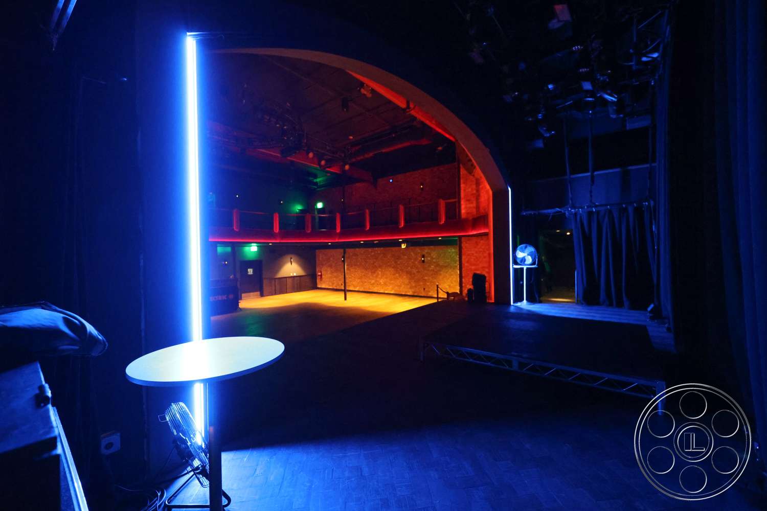 Club 74 - exposed brick walls, dark wood floor, industrial ceiling fixtures, theater stage lighting, arched theater entrance, red balcony railing, LED neon lighting features, sliding theater curtain, ambient stage illumination