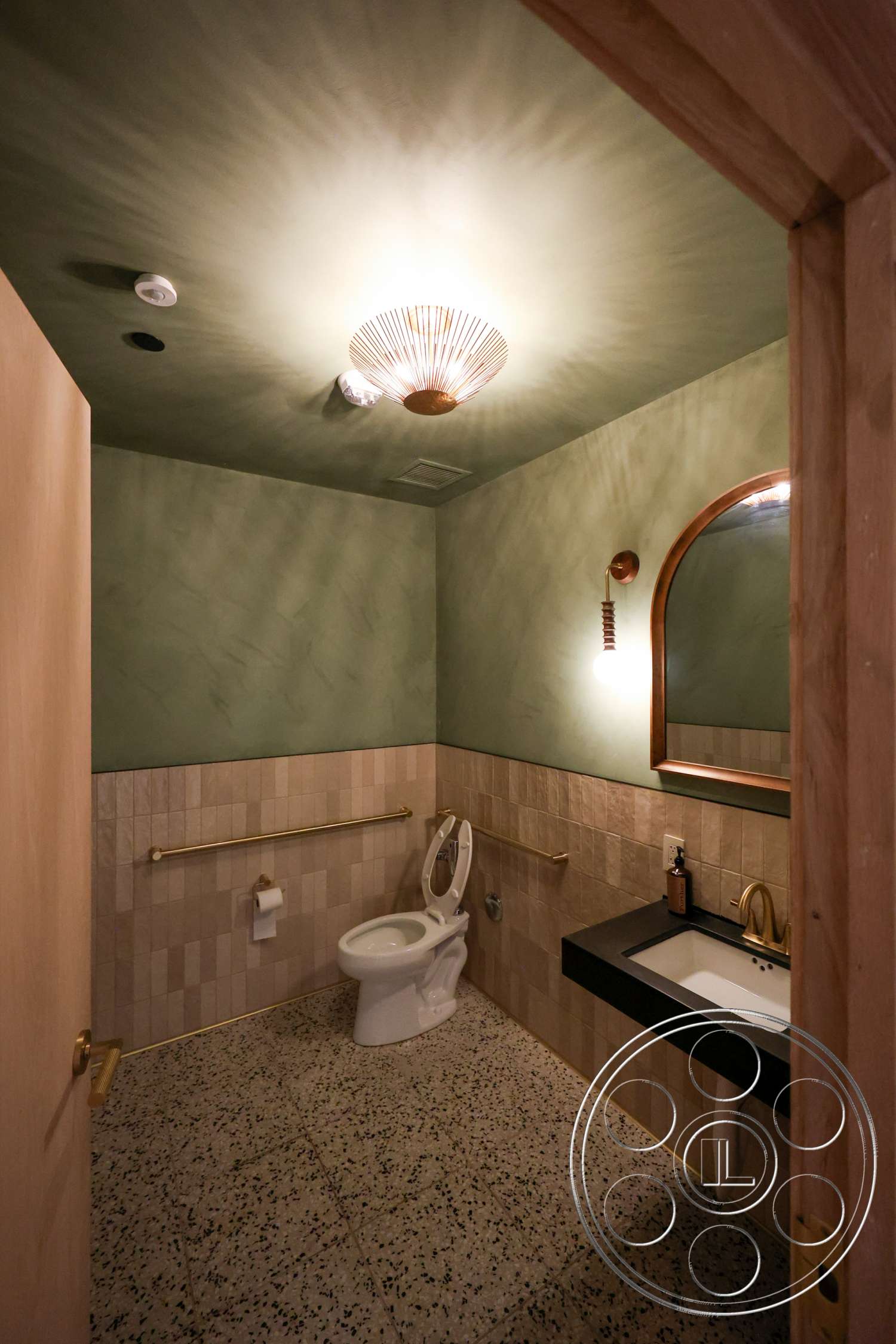 Club 74 - textured plaster walls, polished terrazzo flooring, ceiling light fixture, wooden door frame, beige tiled bathroom walls, round mirror with wooden frame, black composite sink, gold metal faucet, gold grab bars