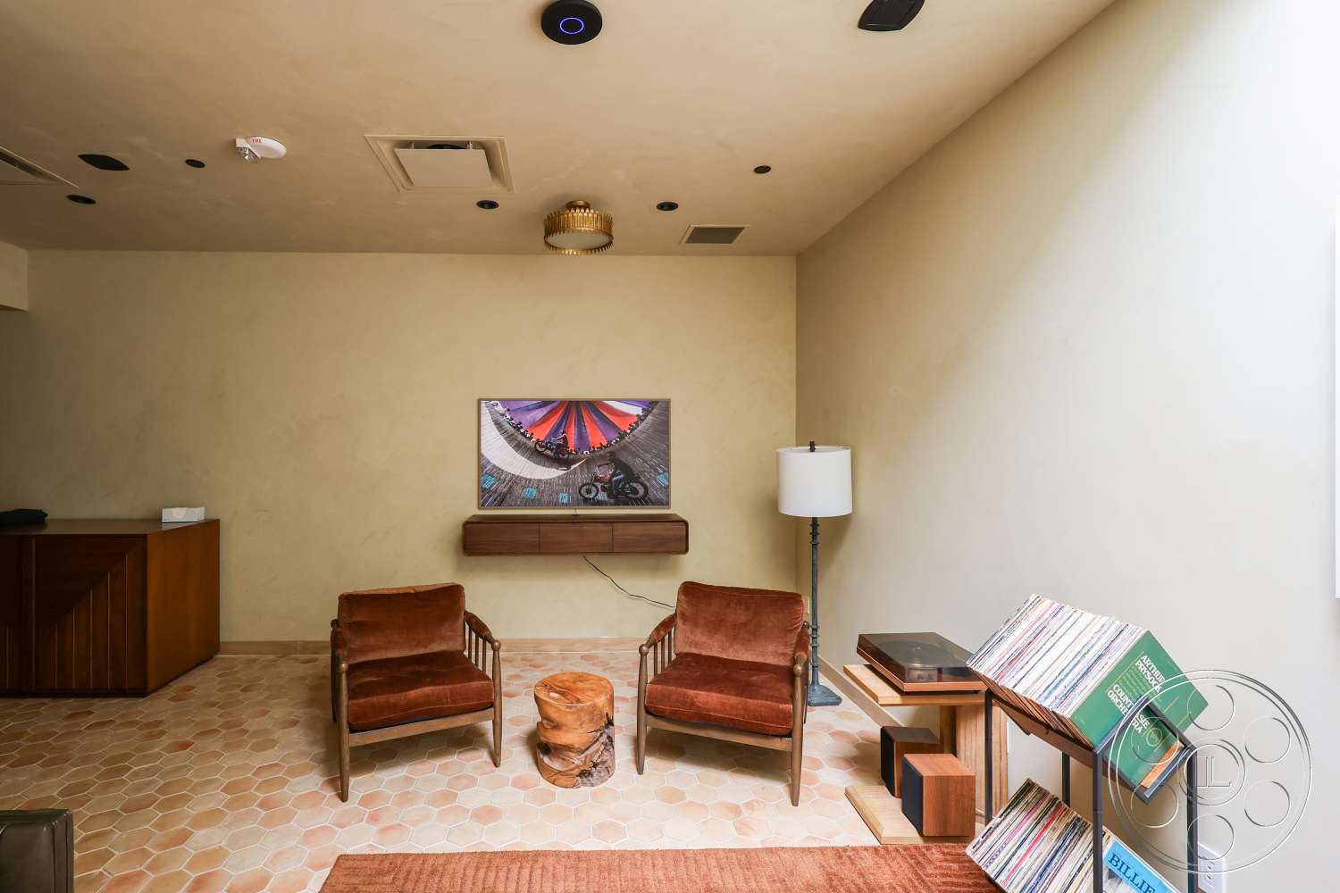 Club 74 - decorative area rug, wooden side table, textured plaster walls, flat-screen television, hexagonal tile flooring, modern ceiling light fixture, brown velvet armchairs, wooden record shelf, minimalist media cabinet