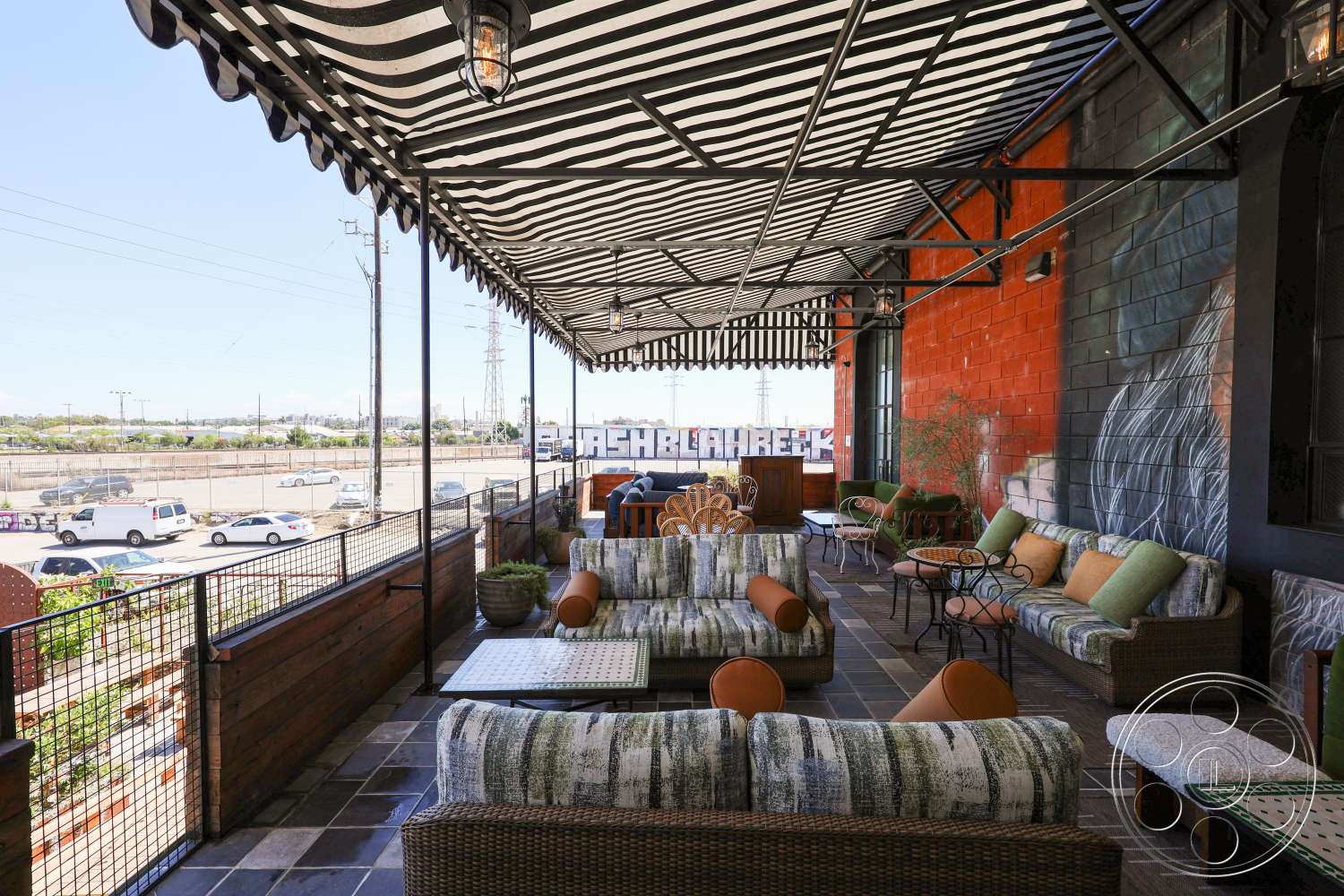 Club 74 - wooden railing, striped awning, decorative outdoor seating, woven lounge chairs, stone tile flooring, exterior patio area, metal railing details, urban setting view, mural accent wall, outdoor coffee tables, green cushion accents