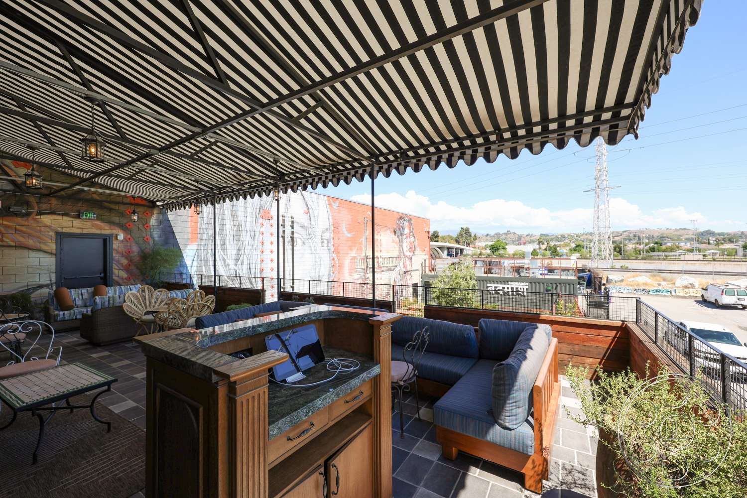 Club 74 - concrete flooring, landscaped planters, stone countertop, decorative lanterns, wooden bar counter, striped awning, exterior patio area, surrounding city view, metal outdoor table, woven rattan furniture, urban mural backdrop, open-air sitting area