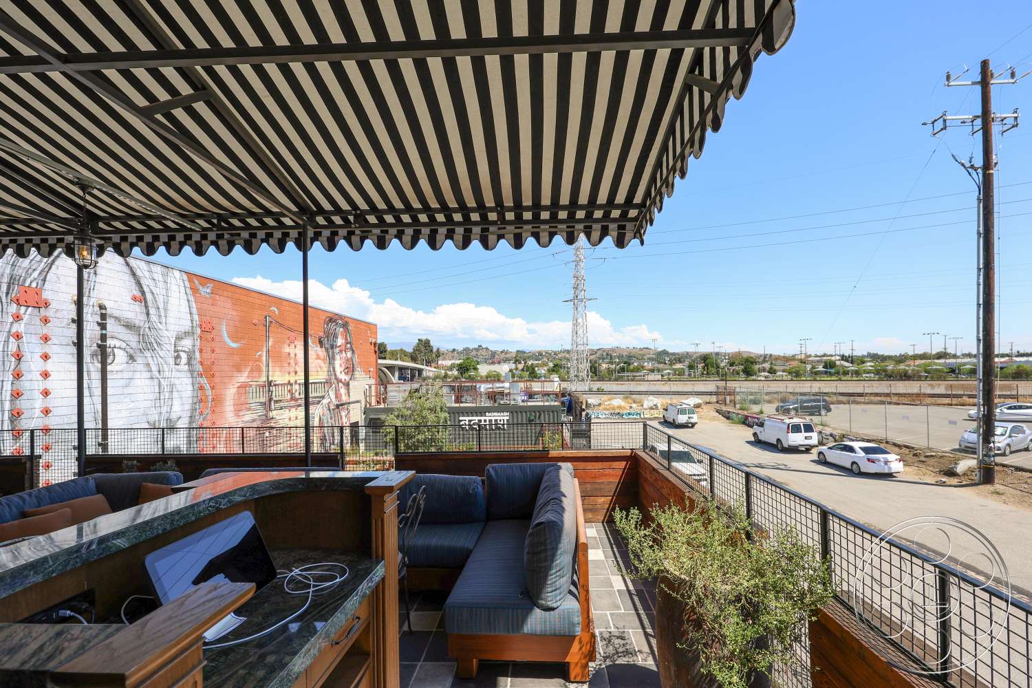 Club 74 - exterior, city view, wooden deck, metal railing, urban setting, striped awning, tile patio flooring, curved outdoor seating, landscaping with greenery, stone bar countertop, exposed brick wall mural