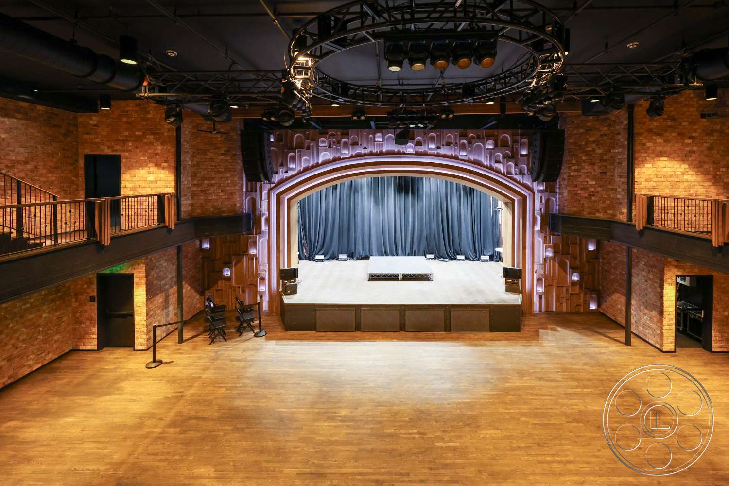 Club 74 - exposed brick walls, wooden floorboards, metal lighting rig, wooden stage flooring, wooden balcony railing, stage with black curtain backdrop, arch-shaped proscenium arch, black ceiling tiles, modern auditorium