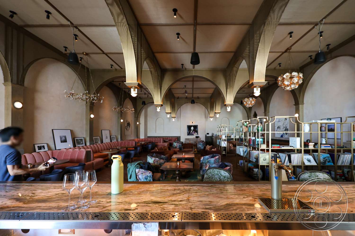 Club 74 - high ceilings, wooden flooring, exposed wooden beams, metal shelving units, framed wall art, textured plaster walls, ambient pendant lighting, marble bar countertop, arched wall forms, upholstered pink banquette seating, velvet armchairs, striped decorative pillows, decorative chandeliers, open lounge area, modern café style