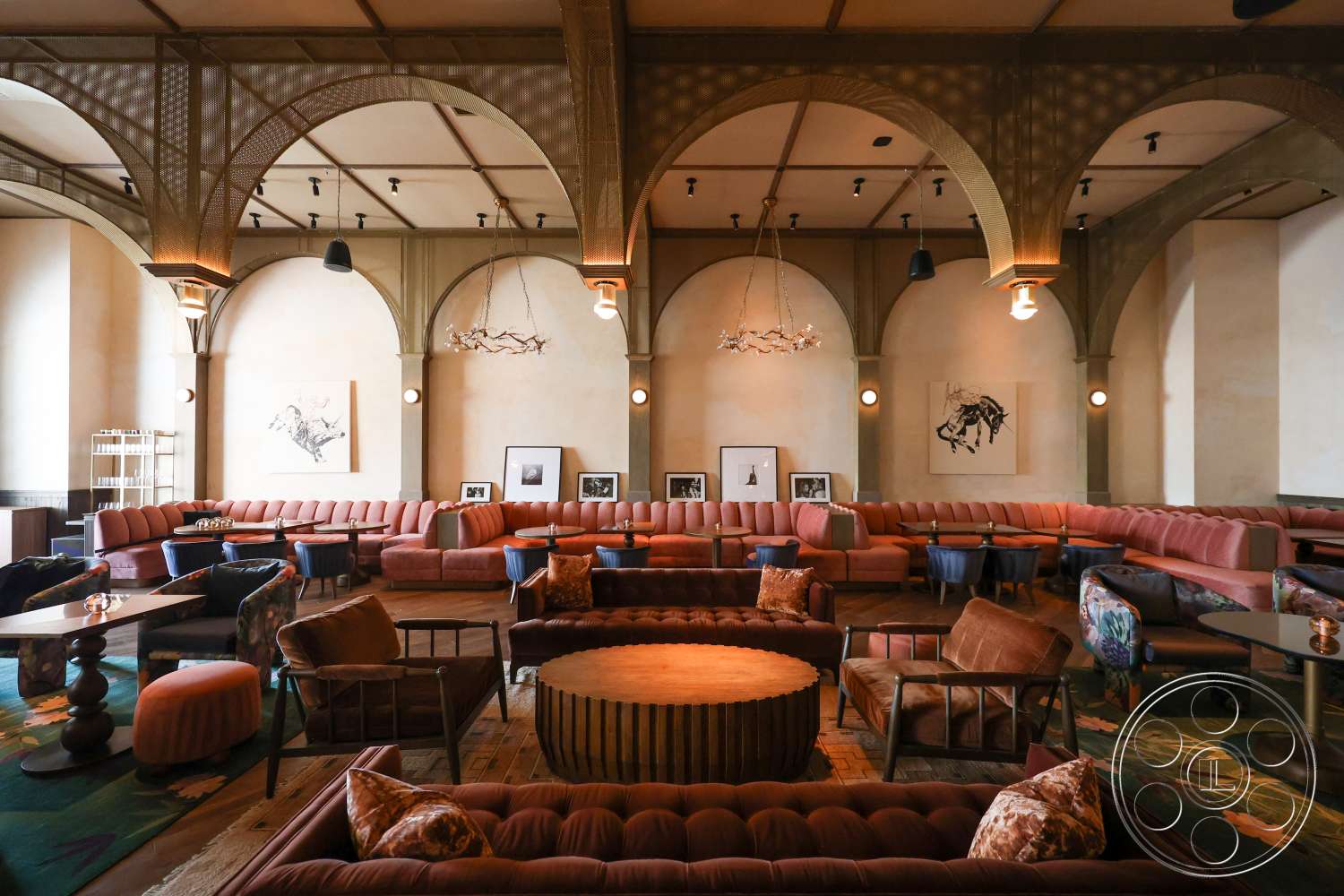 Club 74 - artwork on walls, soft ambient lighting, circular wooden coffee table, textured plaster walls, bar area, large sectional sofa, chandelier light fixture, arched ceiling details, wooden coffee tables, vintage-style light fixtures, dark wood panel flooring, colorful upholstered armchairs, sculptural ceiling elements, open lounge space