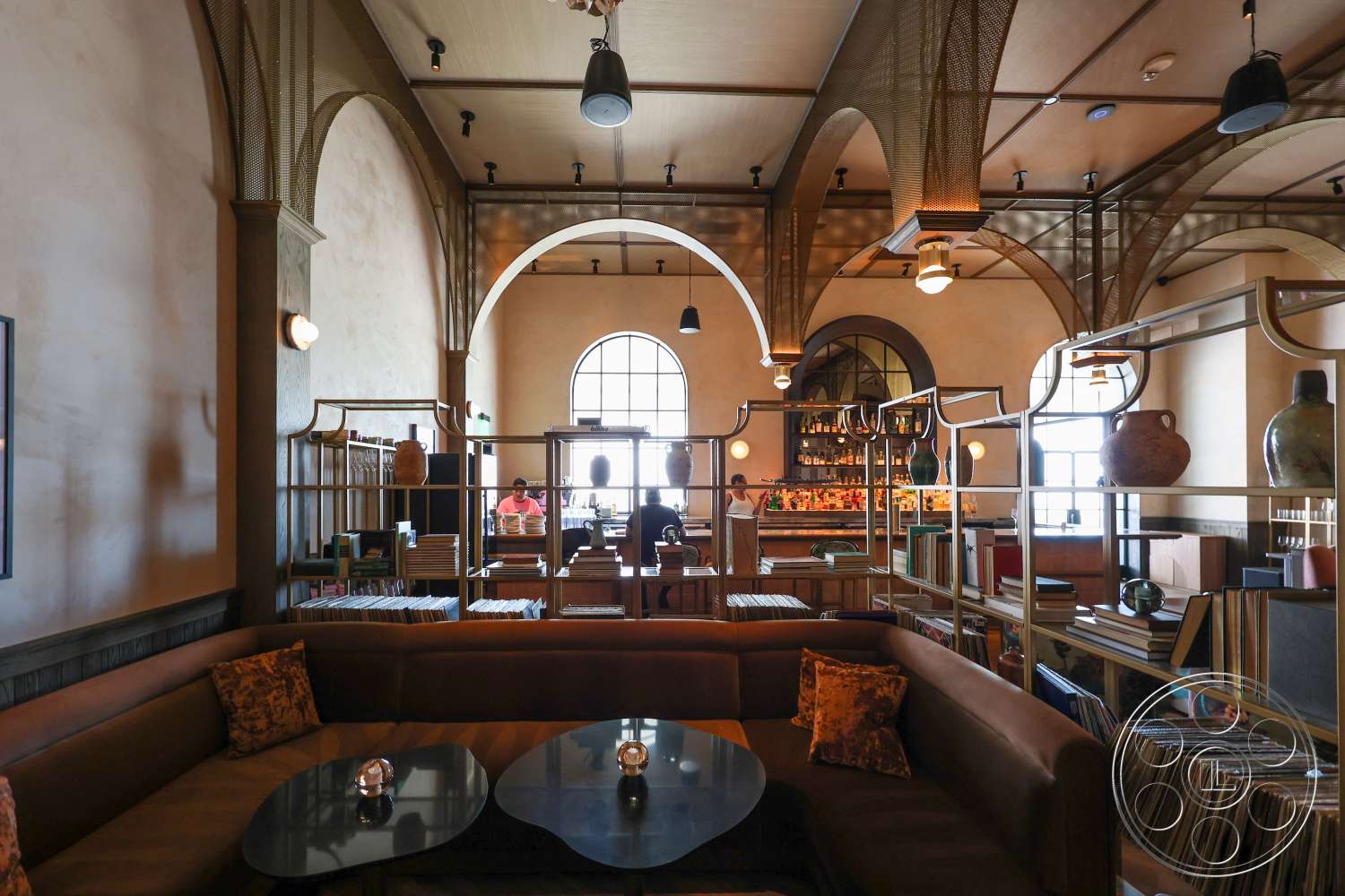 Club 74 - arched windows, plaster wall finish, wooden accent wall, round side tables, modern bar area, upholstered sectional sofa, high ceiling with beams, geometric shapes in furniture, light fixtures with metal shades, metal bookcase dividers, contemporary lounge setting