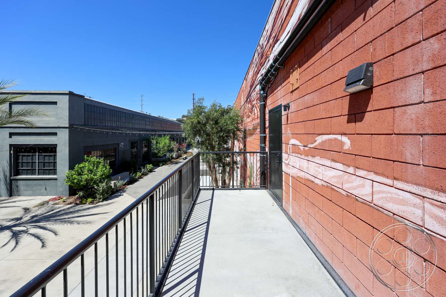 Club 74 - exterior, palm trees, landscaped greenery, urban setting, large industrial windows, clear blue sky, metal balcony railing, concrete balcony floor, textured red block wall, gray industrial building
