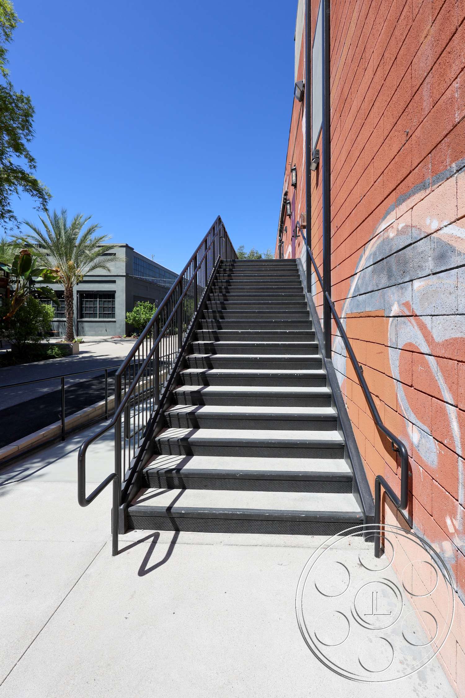 Club 74 - exterior, urban setting, paved walkway, graffiti wall art, industrial-style architecture, metal stair railings, painted concrete walls, textured concrete steps, concrete staircase, palm trees in foreground