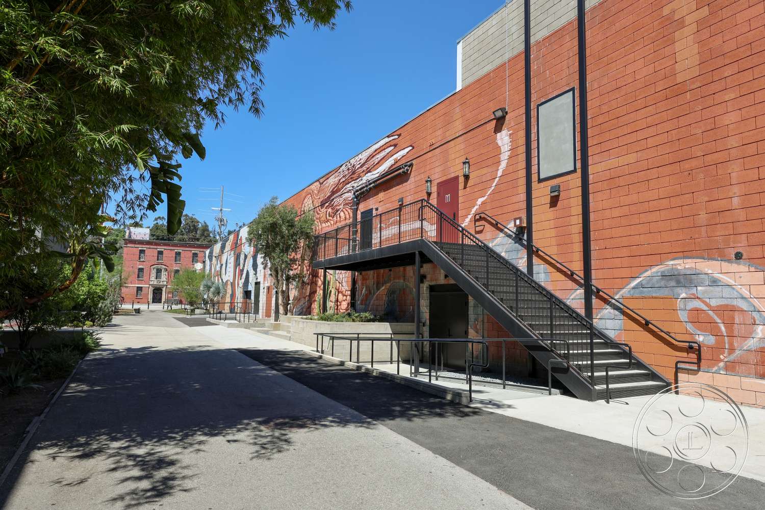 Club 74 - exterior, landscaped greenery, concrete pathway, urban setting, textured plaster wall, industrial architectural style, open courtyard layout, brick wall with mural, metal fire escape staircase, multi-story brick building