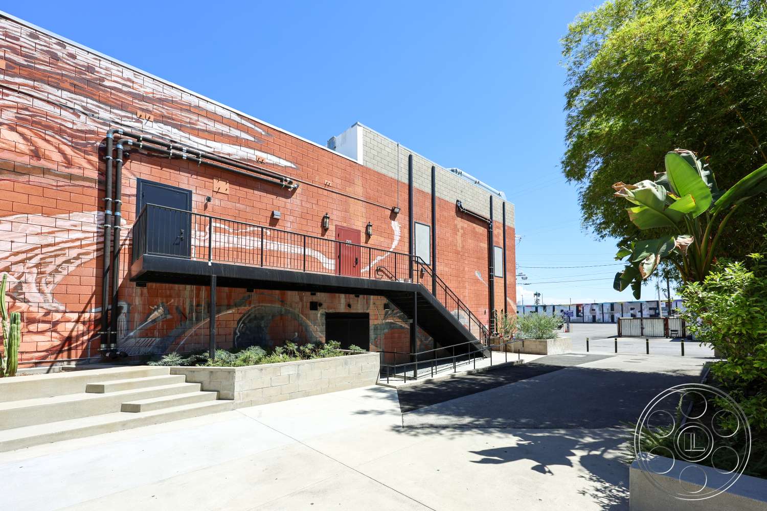 Club 74 - exterior, metal railing, gray concrete wall, urban setting, concrete steps, clear blue sky, black metal staircase, modern industrial design, landscaped garden beds, concrete patio floor, brick wall with mural, horizontal metal pipes