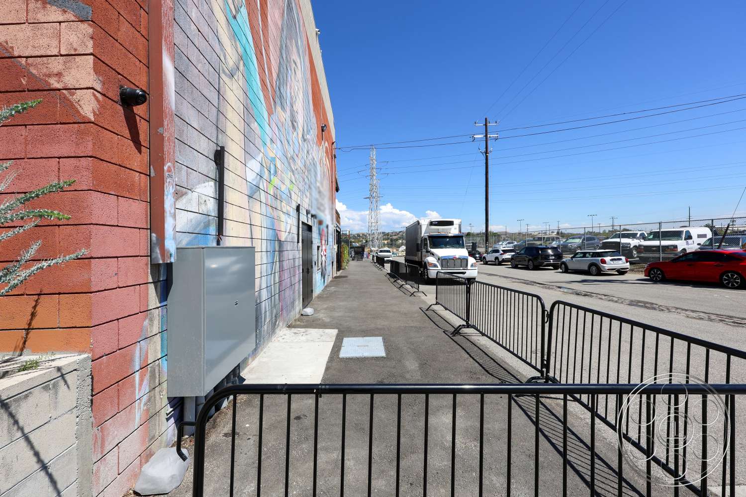 Club 74 - exterior, urban setting, blue sky, utility poles, asphalt parking lot, concrete sidewalk, painted brick wall, mural detail on wall, metal electrical box, metal barrier fencing