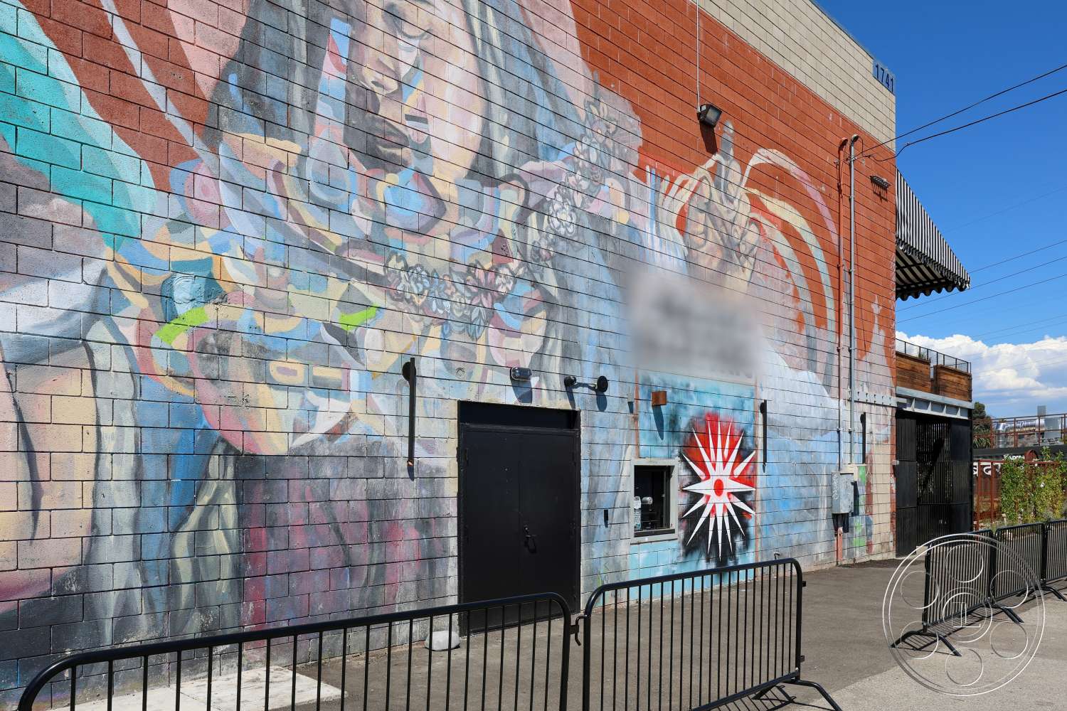 Club 74 - exterior, outdoor seating area, urban setting, paved surface, chain link fence, industrial style architecture, metal awning, cinder block walls, multi-colored mural, black steel entrance door