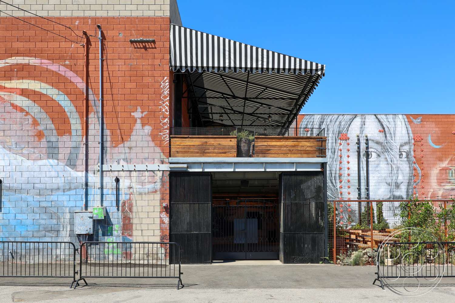 Club 74 - exterior, concrete flooring, urban setting, exposed brick wall, industrial design elements, metal gates, landscaped patio area, wooden balcony railing, colorful mural artwork, metal structural beams, striped canopy awning