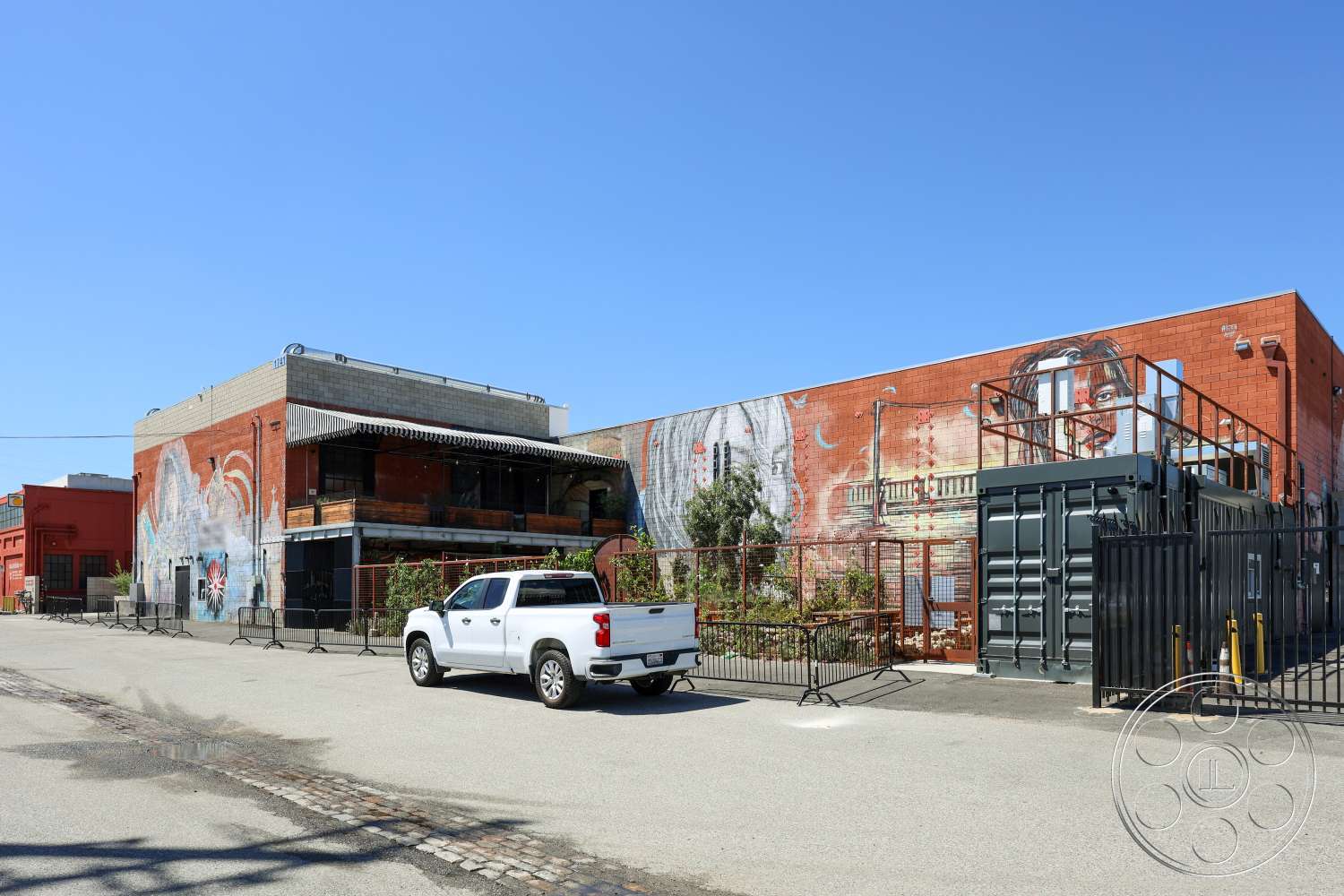 Club 74 - exterior, concrete floor, urban setting, metal fencing, steel staircase, industrial style architecture, brick building facade, colorful mural artwork, wraparound balcony, black shipping container, cobblestone street