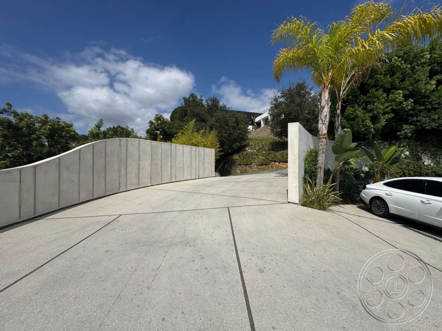 The Bellagio Estate - exterior, modern architectural style, tropical palm trees, curved concrete driveway, landscaped hillside setting, concrete privacy wall, bushes and greenery, textured concrete driveway surface, concrete gate
