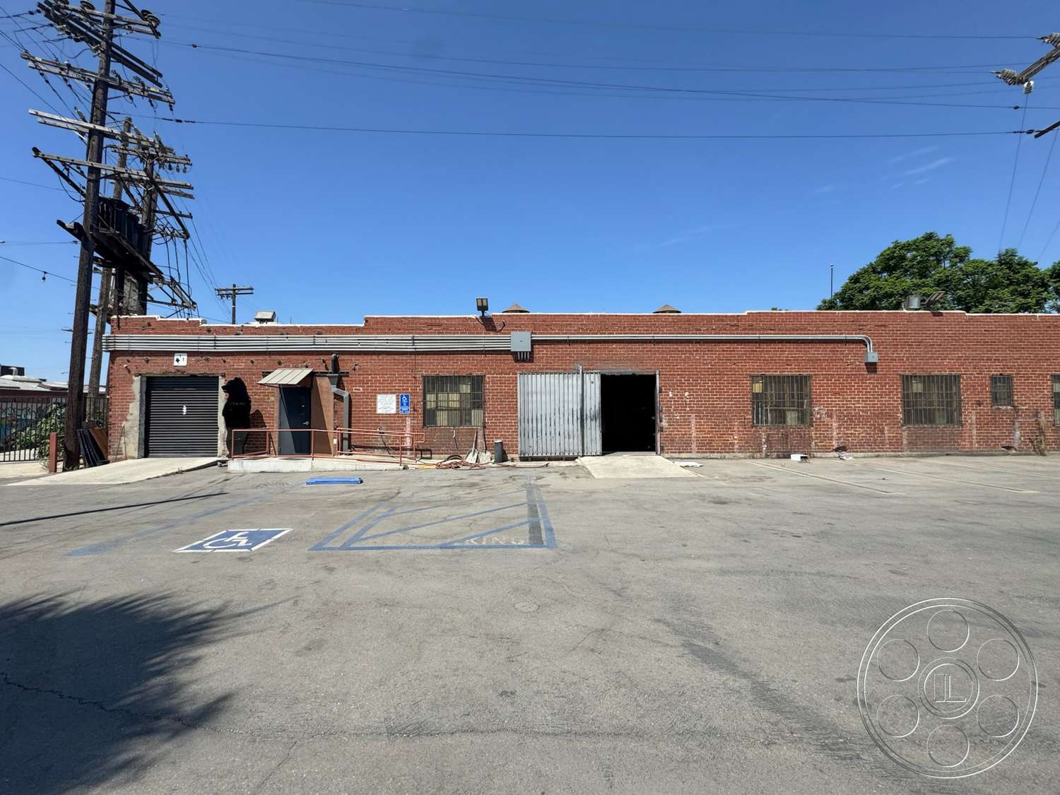 Warehouse 62 - exterior, paved driveway, concrete parking lot, industrial setting, overhead power lines, flat roof structure, metal garage door, red brick warehouse wall, large open entrance, steel window bars, sidewalk access ramp