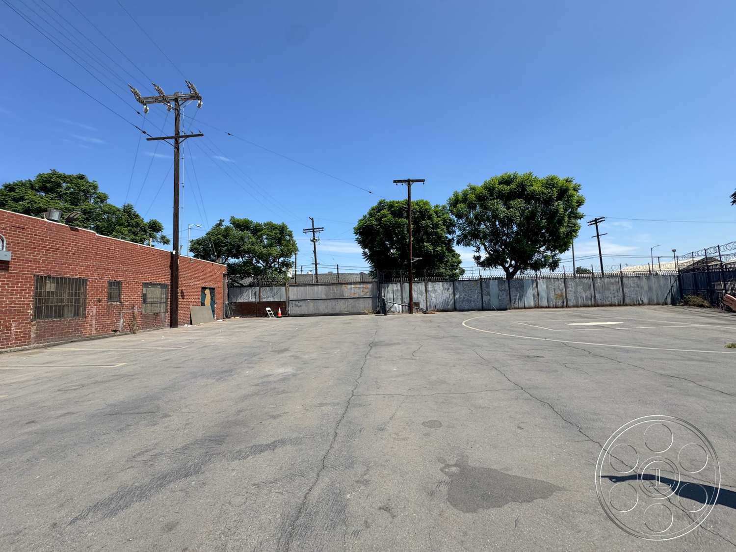 Warehouse 62 - exterior, mature trees, clear blue sky, chain link fence, asphalt parking lot, telephone poles, red brick wall, industrial setting, flat landscape