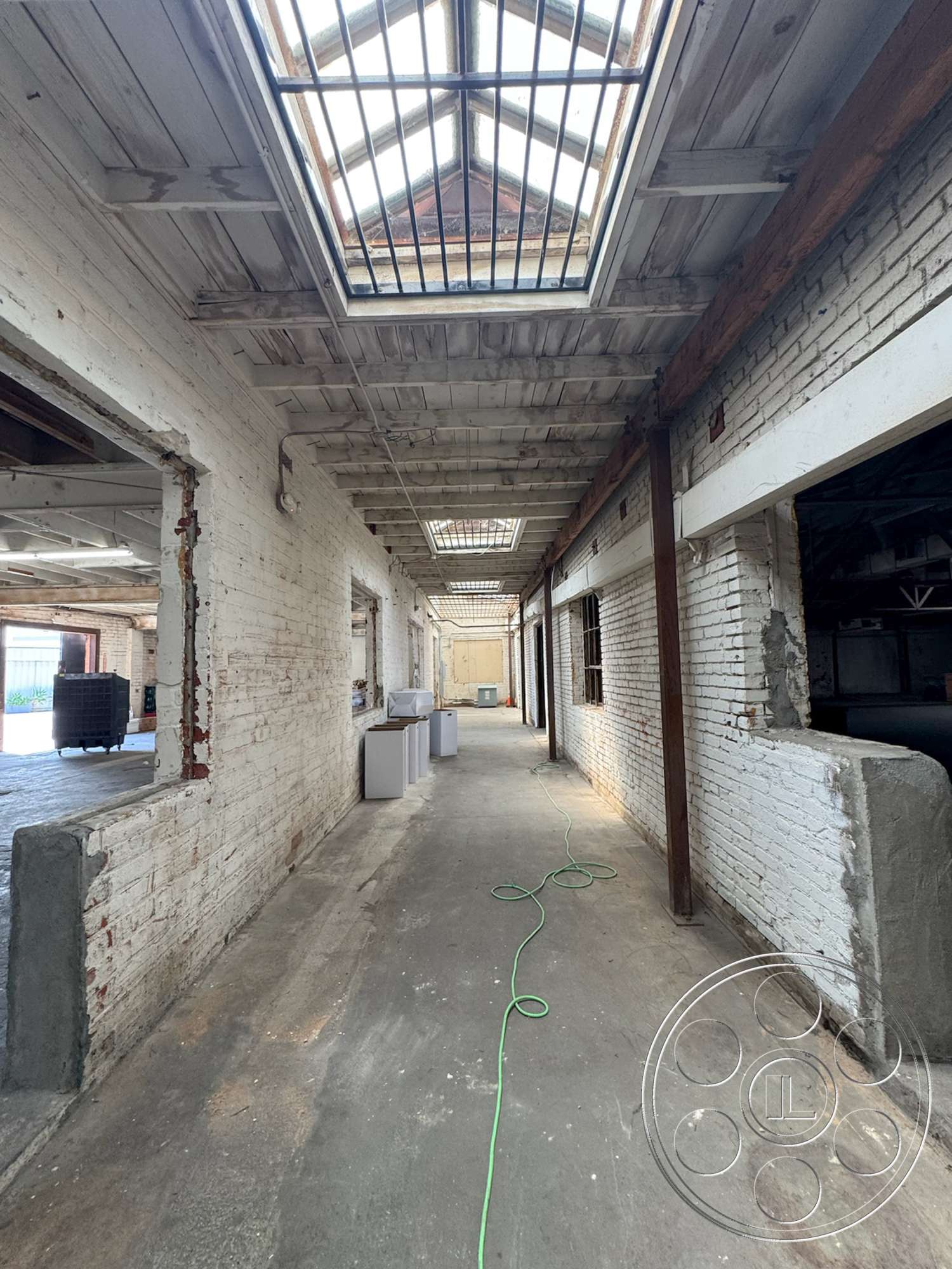 Warehouse 62 - concrete flooring, exposed brick walls, interior hallway, wood beam ceiling, industrial corridor, skylights with metal framing, oak wood door frames, wiring along the floor, whitewashed brick texture