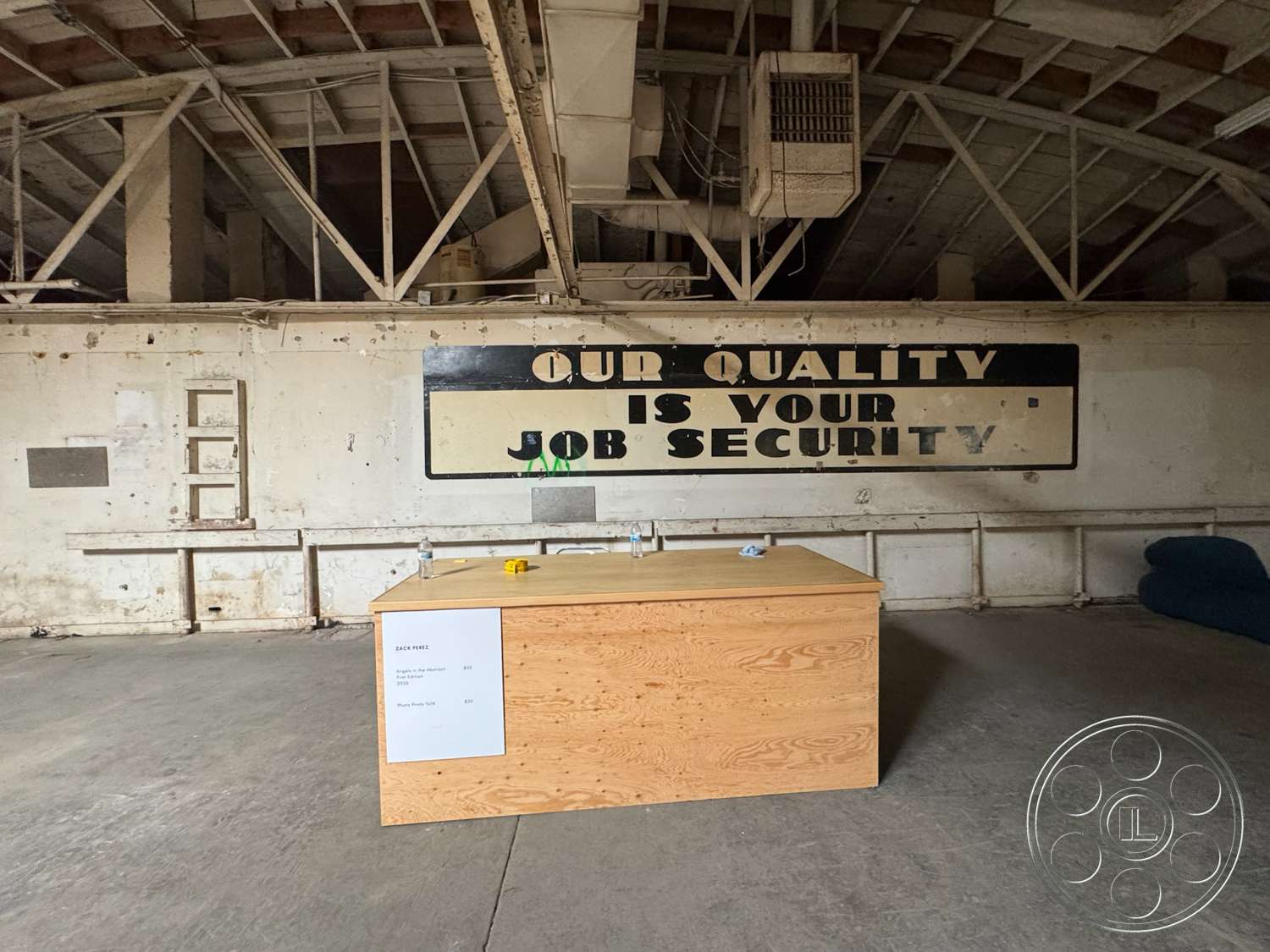 Warehouse 62 - open floor plan, concrete floor, metal shelving unit, dimly lit space, plain white painted walls, exposed wooden beam ceiling, large industrial wall-mounted fan, wooden countertop island, historic building details, vintage wall sign, urban warehouse environment