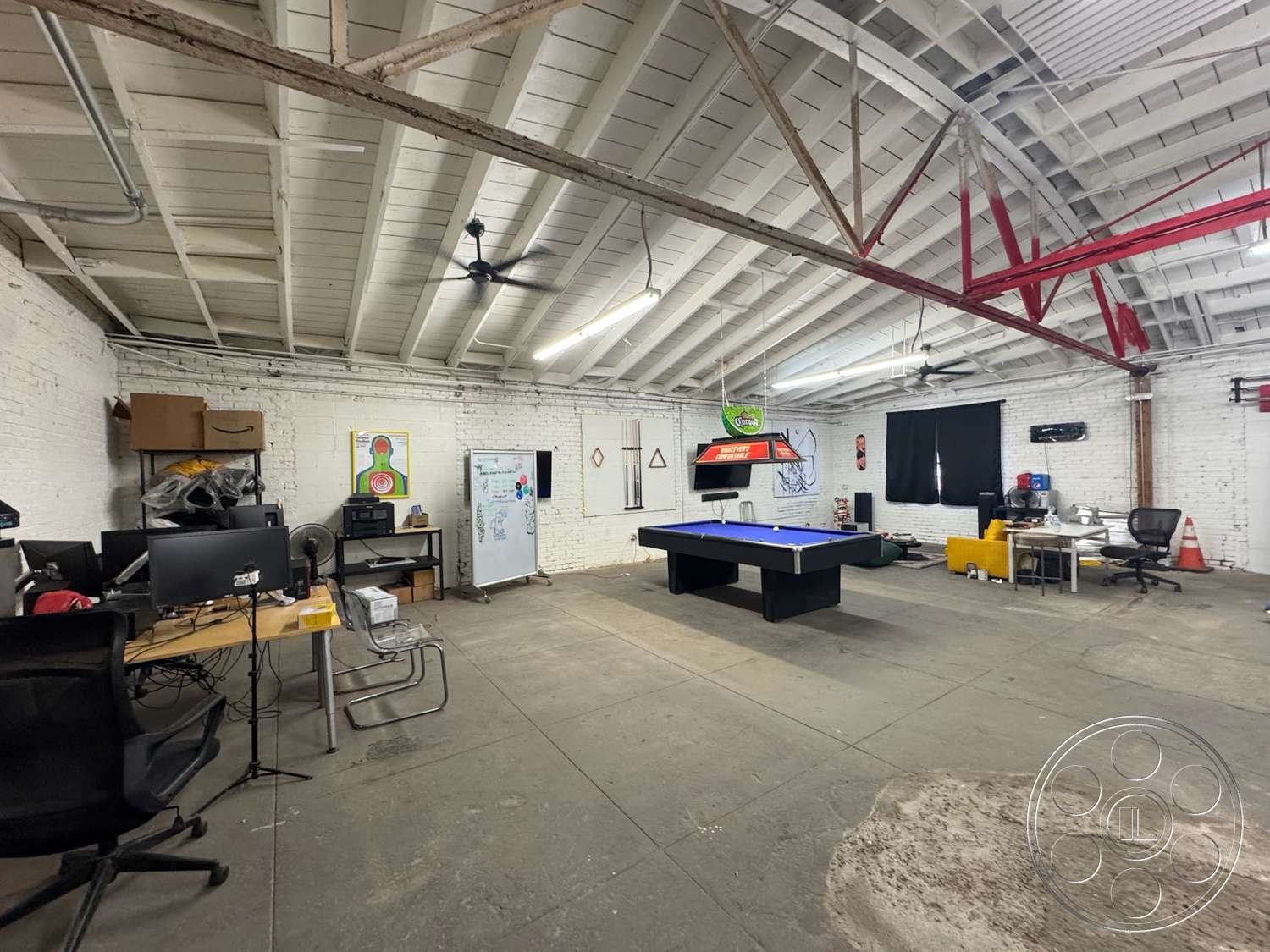 Warehouse 62 - concrete floor, pool table, exposed wood ceiling beams, industrial style interior, bright lighting fixtures, black ergonomic office chair, white painted brick walls, steel office desk, fan hanging from ceiling, large windows with black curtains, workspace with wooden table, urban garage setting