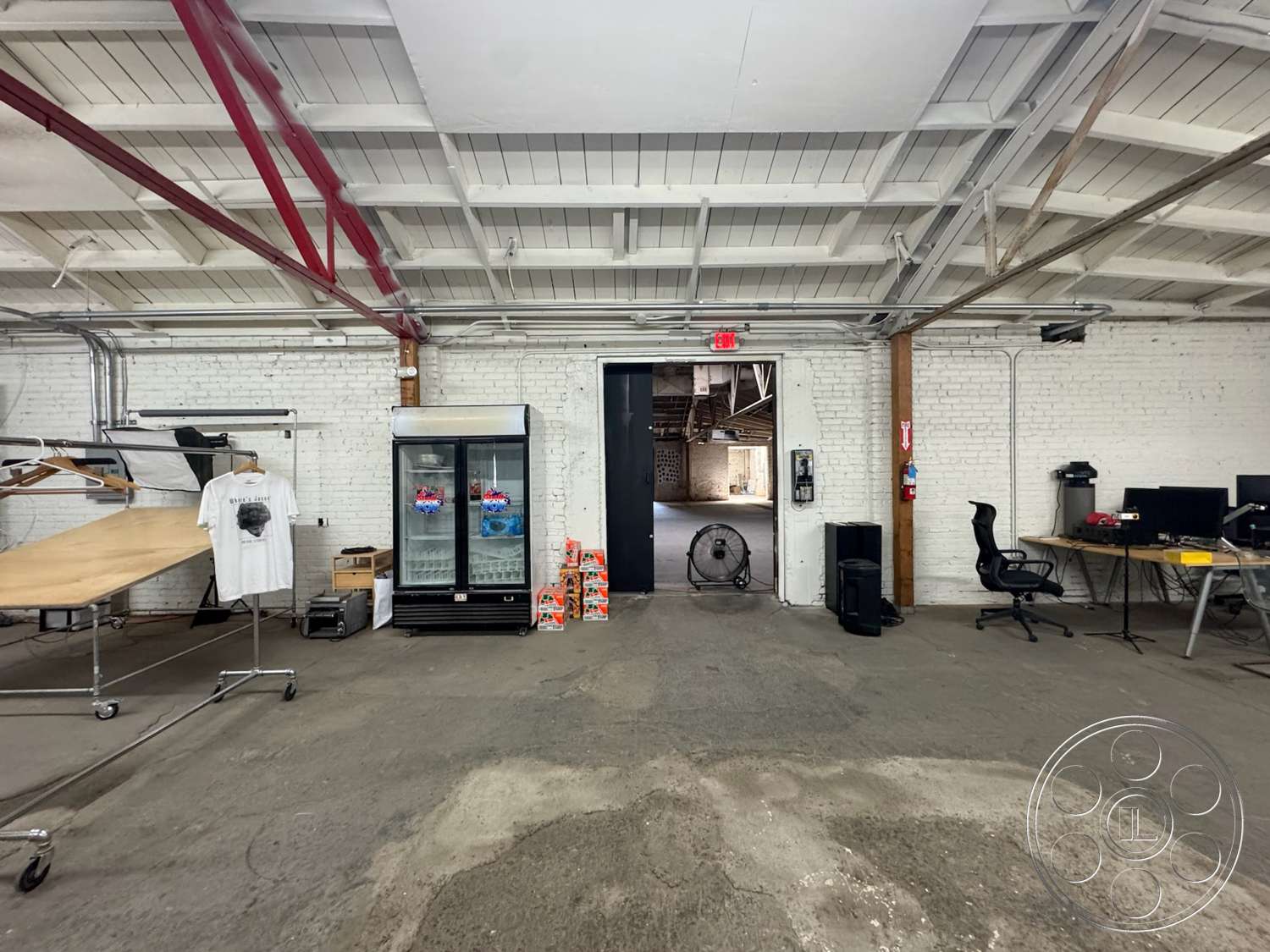 Warehouse 62 - open floor plan, concrete flooring, exposed brick walls, vaulted ceiling, wooden table, wood beam ceiling, industrial style interior, metal clothing rack, glass refrigerator unit, red steel support beams, office workstation setup, doorway with black door