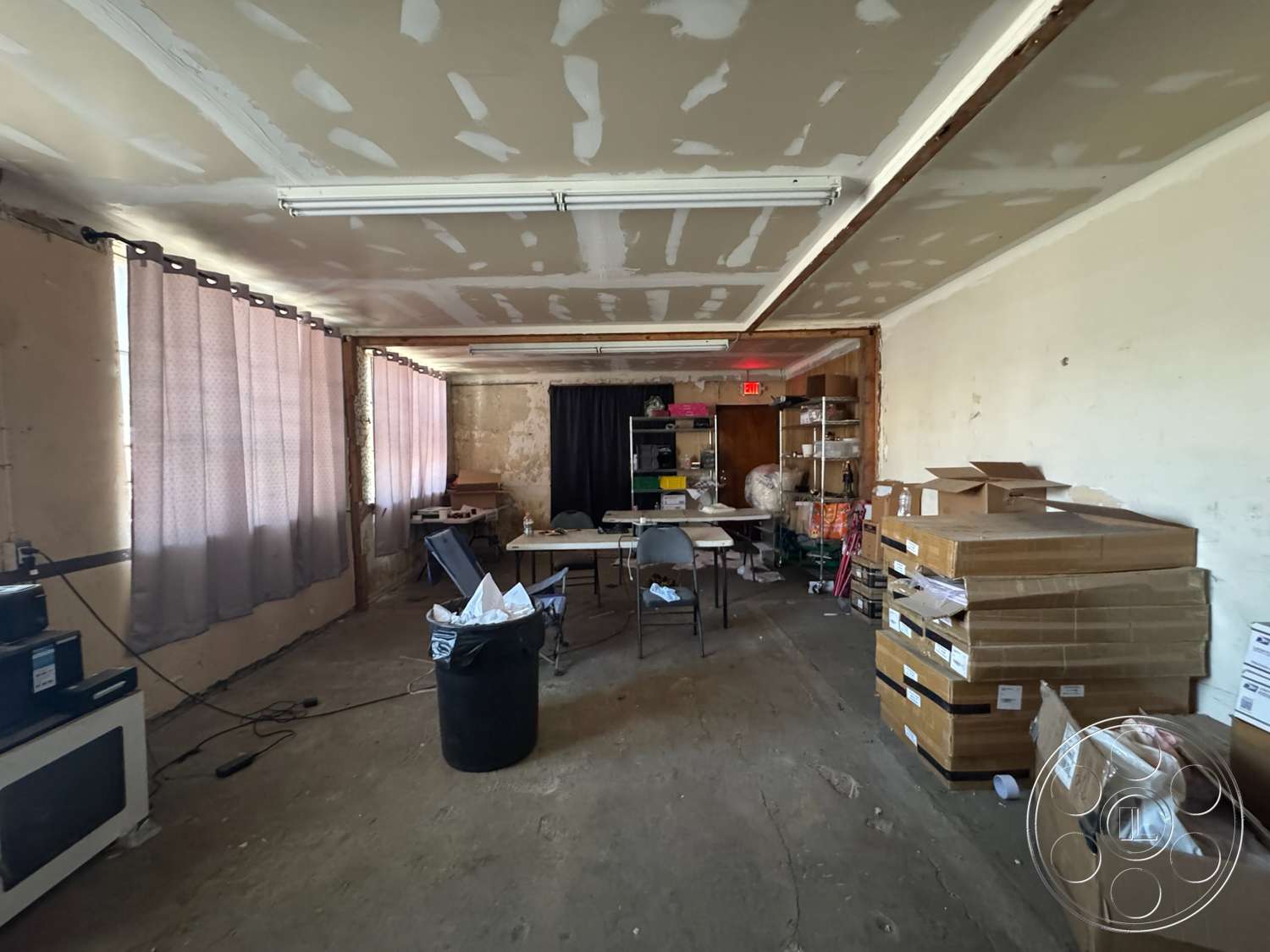 Warehouse 62 - plaster walls, natural light from windows, unfinished concrete floor, window treatments with curtains, drywalled ceiling, open workspace style, storage area with cardboard boxes, basic ceiling light fixtures, simple furniture setup with tables and chairs