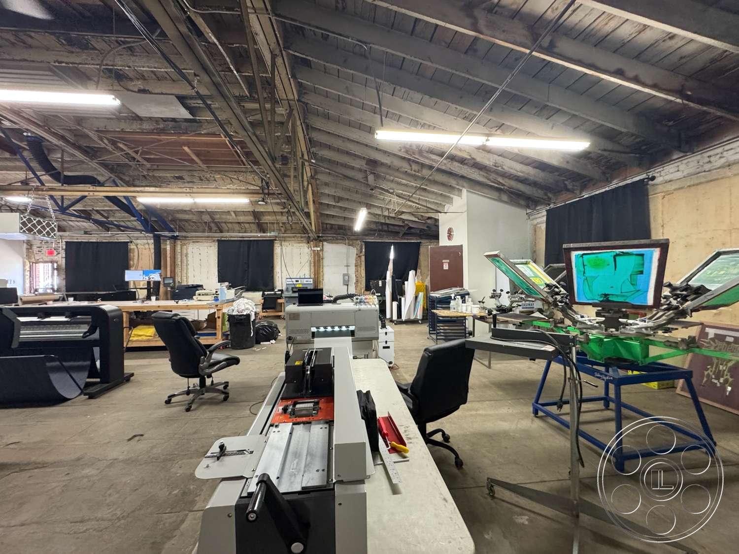 Warehouse 62 - concrete flooring, overhead fluorescent lighting, exposed wooden ceiling beams, metal industrial worktables, steel screen printing equipment, black fabric curtains, light industrial workspace, open layout workshop, blue structural supports