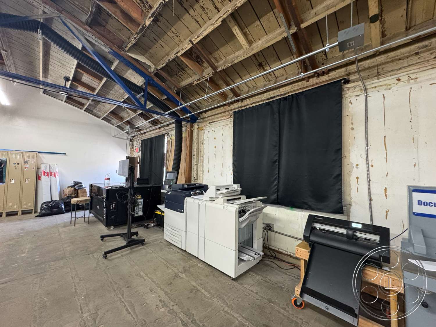 Warehouse 62 - concrete floor, utility space, white painted walls, exposed wooden ceiling beams, open workspace layout, industrial ceiling ductwork, computer workstation, black fabric window treatments, office equipment area, metal file lockers, document printing station, light industrial style