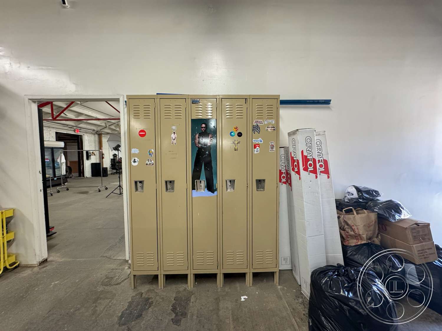 Warehouse 62 - concrete floor, open doorway, bright white walls, exposed ceiling beams, industrial space, cardboard boxes, metal lockers, beige metal locker doors, storage bags, sticker-covered lockers, blue painted beam
