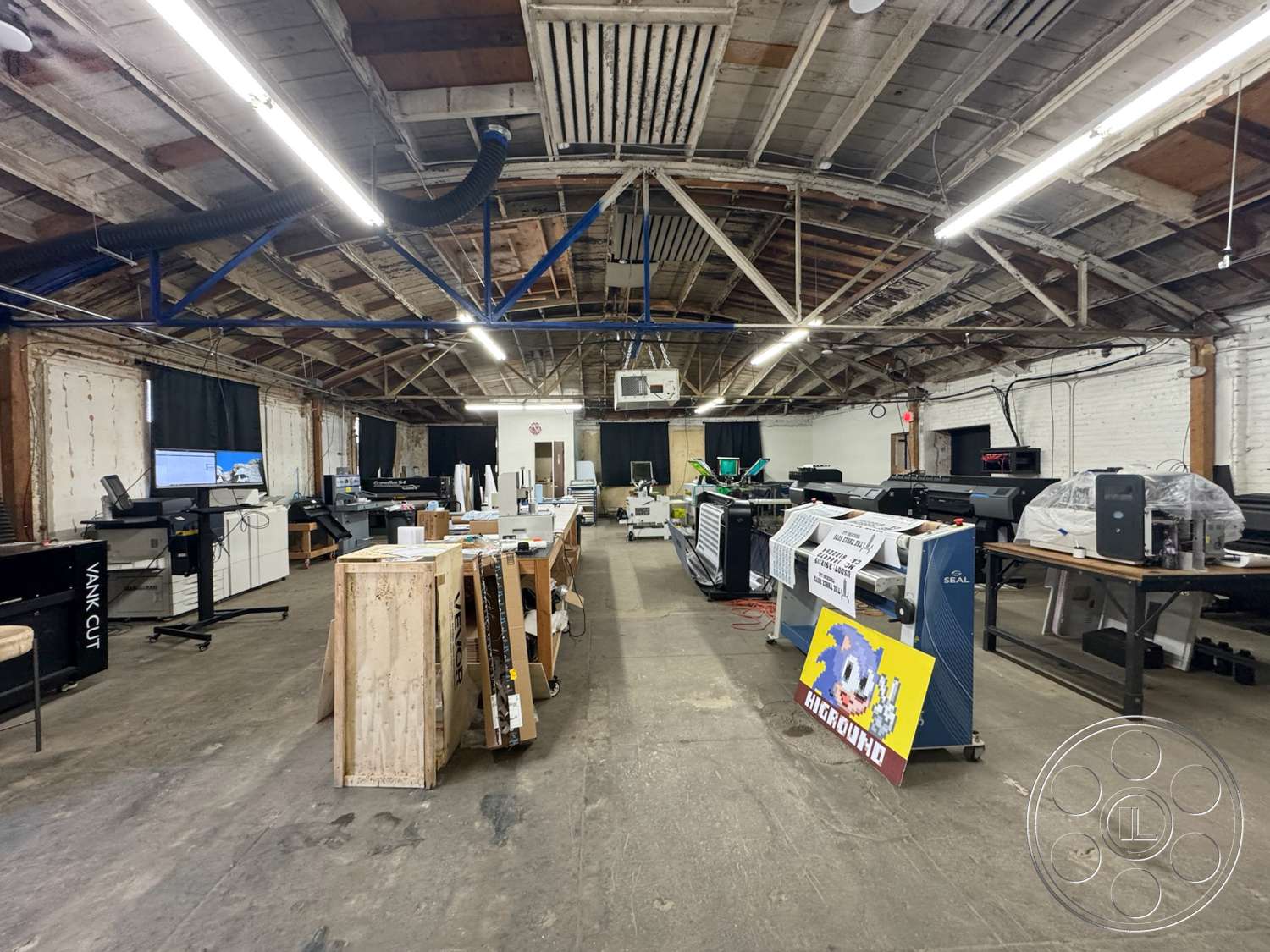 Warehouse 62 - concrete floor, textured plaster walls, exposed wood ceiling beams, functional work environment, industrial ceiling structure, wooden work tables, open workshop space, large-format printers, black-out curtains, workshop lighting, commercial printing area, exterior exposed brick walls