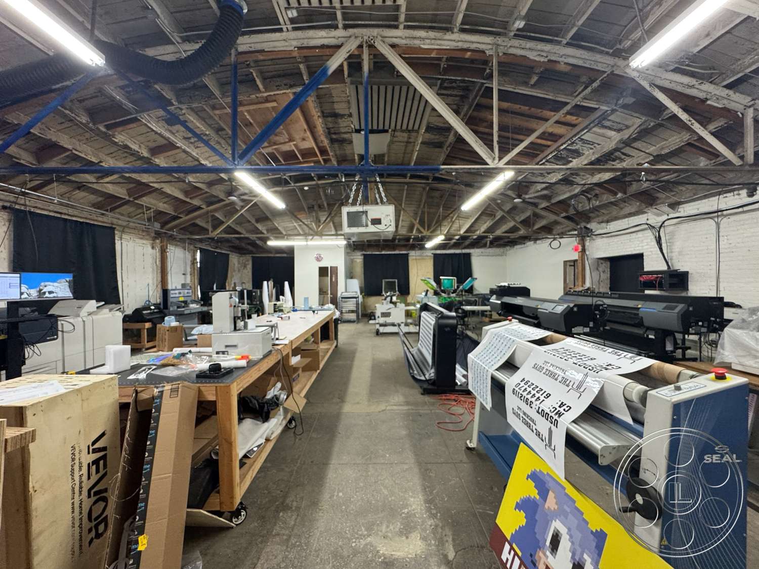 Warehouse 62 - suspended lighting fixtures, ventilation ducts, white painted walls, exposed wooden ceiling beams, open workspace layout, industrial concrete flooring, wooden workshop tables, black backdrop curtains, printing equipment