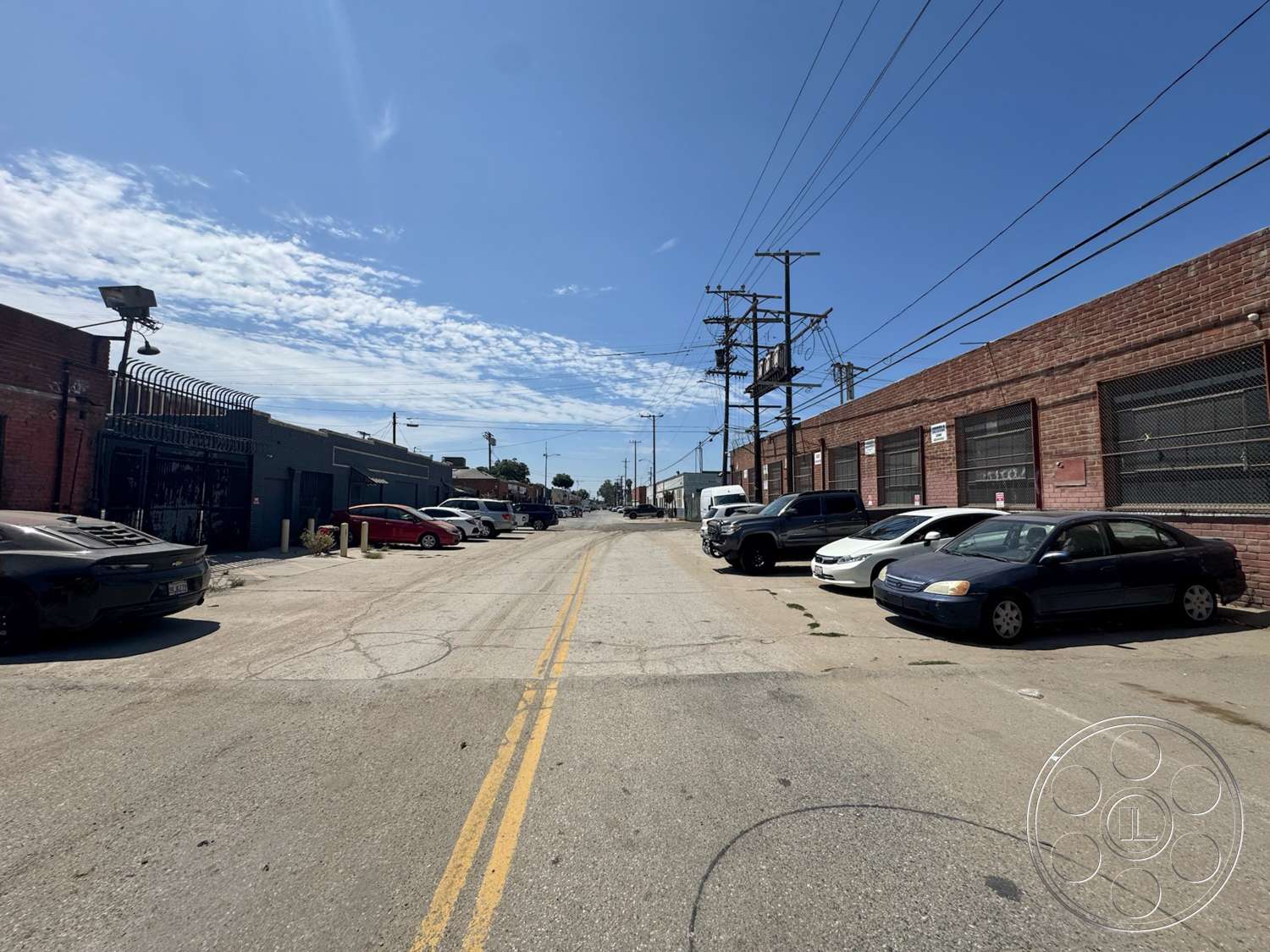 Warehouse 62 - exterior, urban environment, clear blue sky, asphalt road surface, industrial building setting, chain-link fencing, brick warehouse walls, two-lane street, vertical utility poles, vehicle parking on street