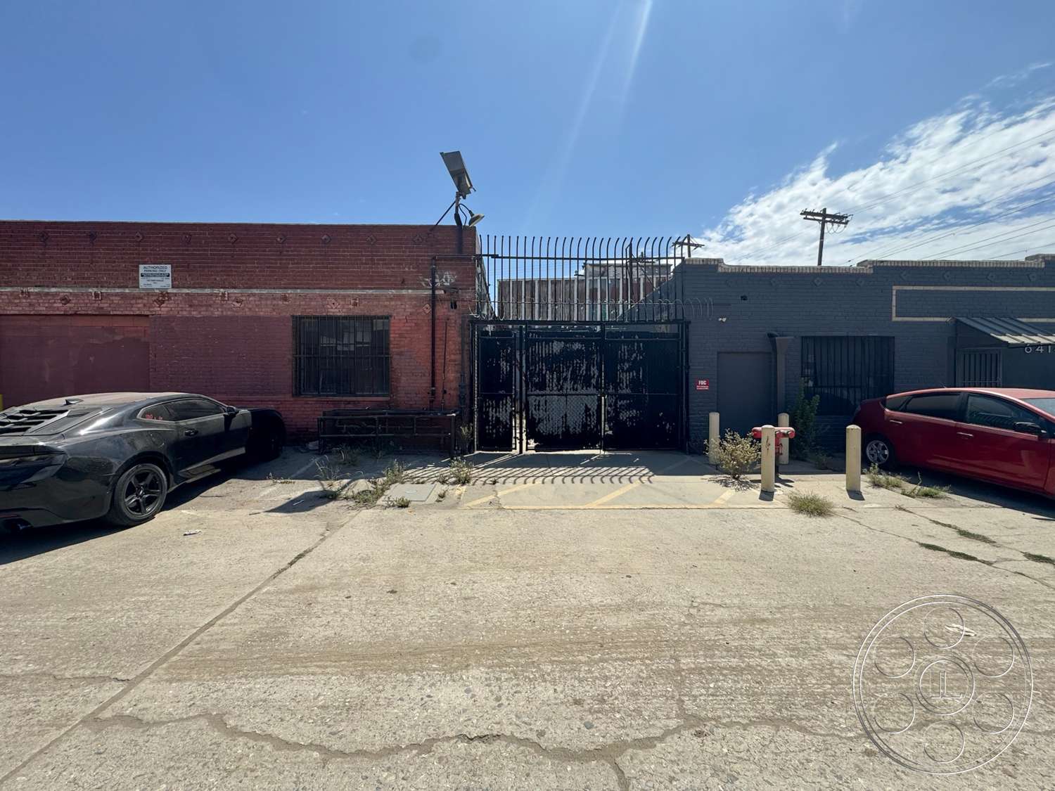 Warehouse 62 - exterior, paved driveway, brick walls, urban environment, black wrought iron fence, industrial setting, red brick facade, overhead utility lines, concrete parking lot surface, metal security gate, window bars, buildings in close proximity, dirt and weeds in pavement cracks