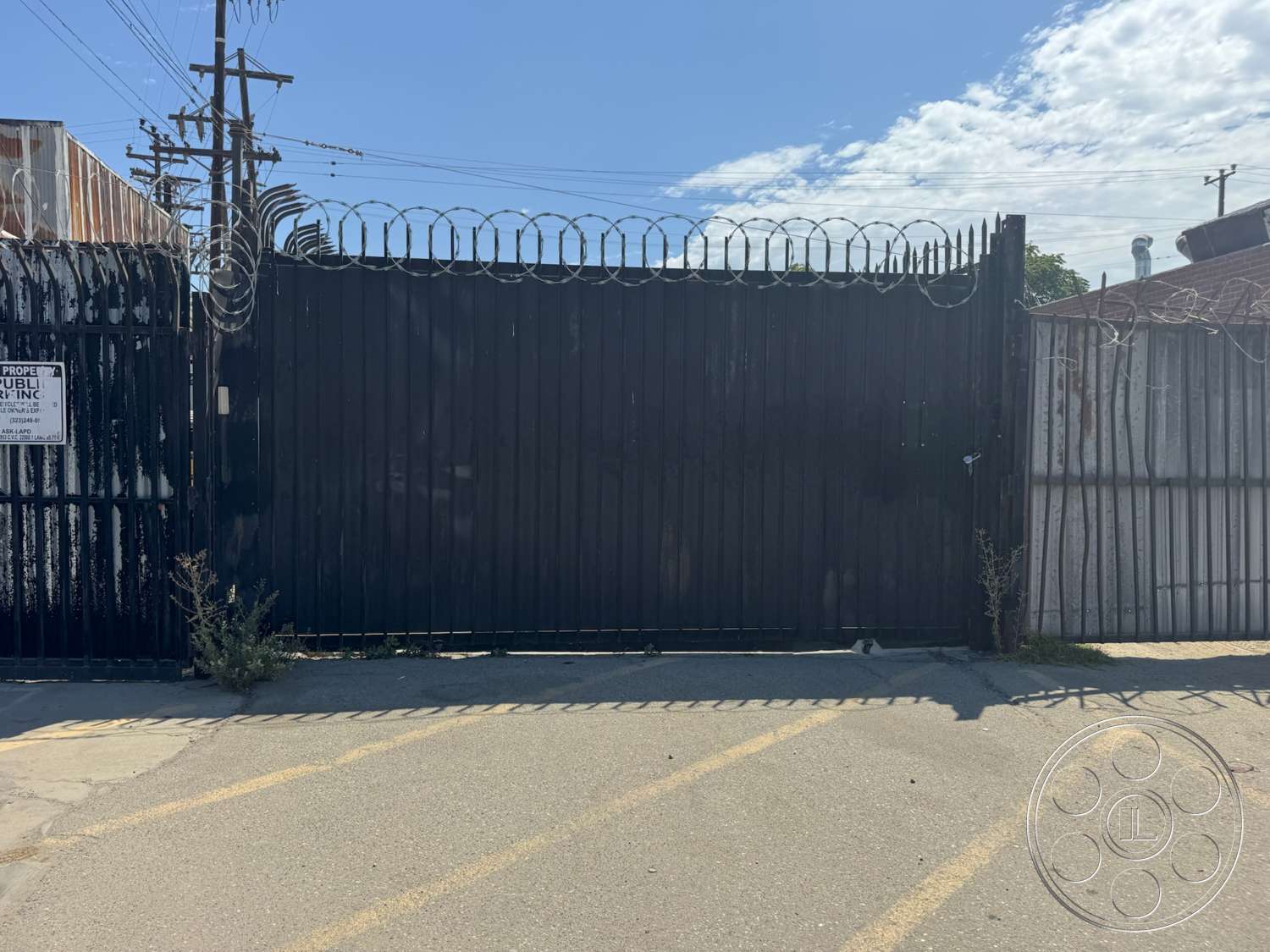 Warehouse 62 - exterior, concrete driveway, overhead power lines, urban industrial setting, black metal security gate, barbed wire top fence, weathered metal building, signage on gate