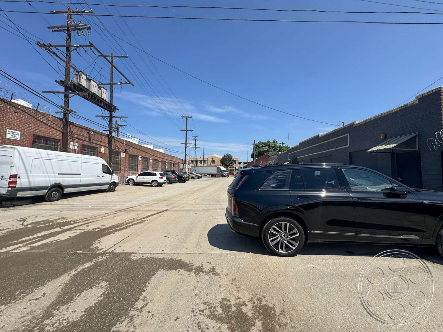 Warehouse 62 - exterior, clear blue sky, parked vehicles, loading area, urban industrial setting, single-story commercial structure, asphalt street surface, brick warehouse building, concrete gravel parking lot, metal overhead garage doors, wooden power poles, utility lines overhead, bicycle rack
