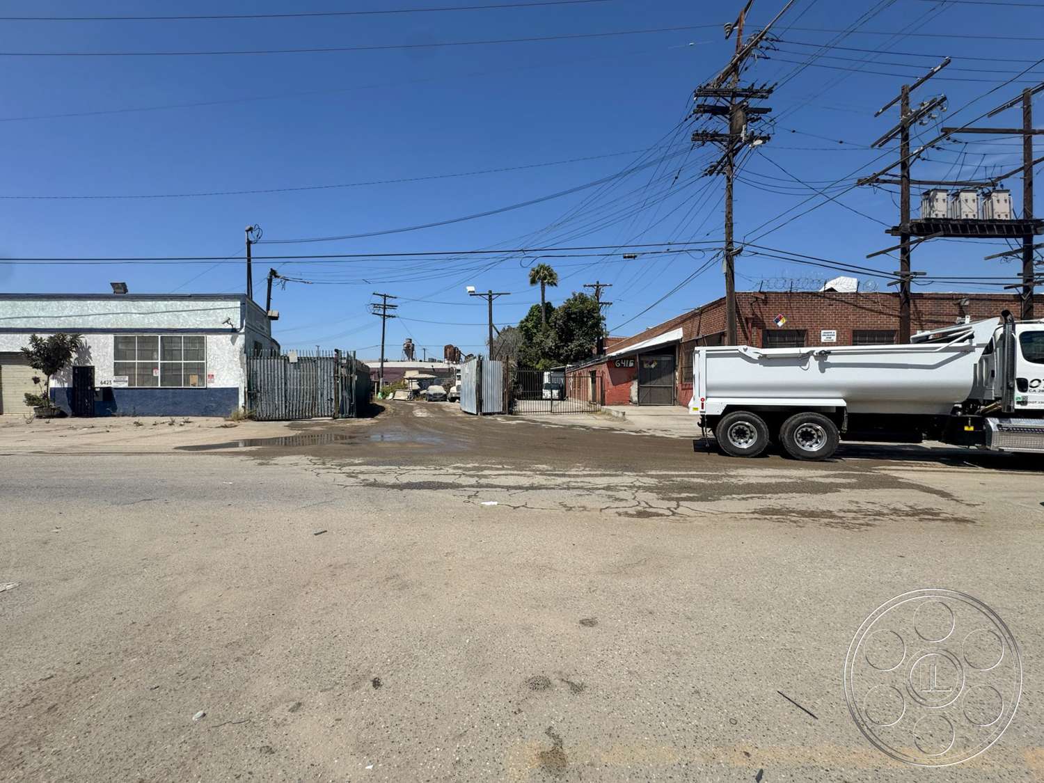 Warehouse 62 - exterior, urban landscape, concrete pavement, metal fence, paved alleyway, gravel ground, utility poles, loading area, industrial setting, overhead power lines, flat roof structure, brick building facade, commercial warehouse