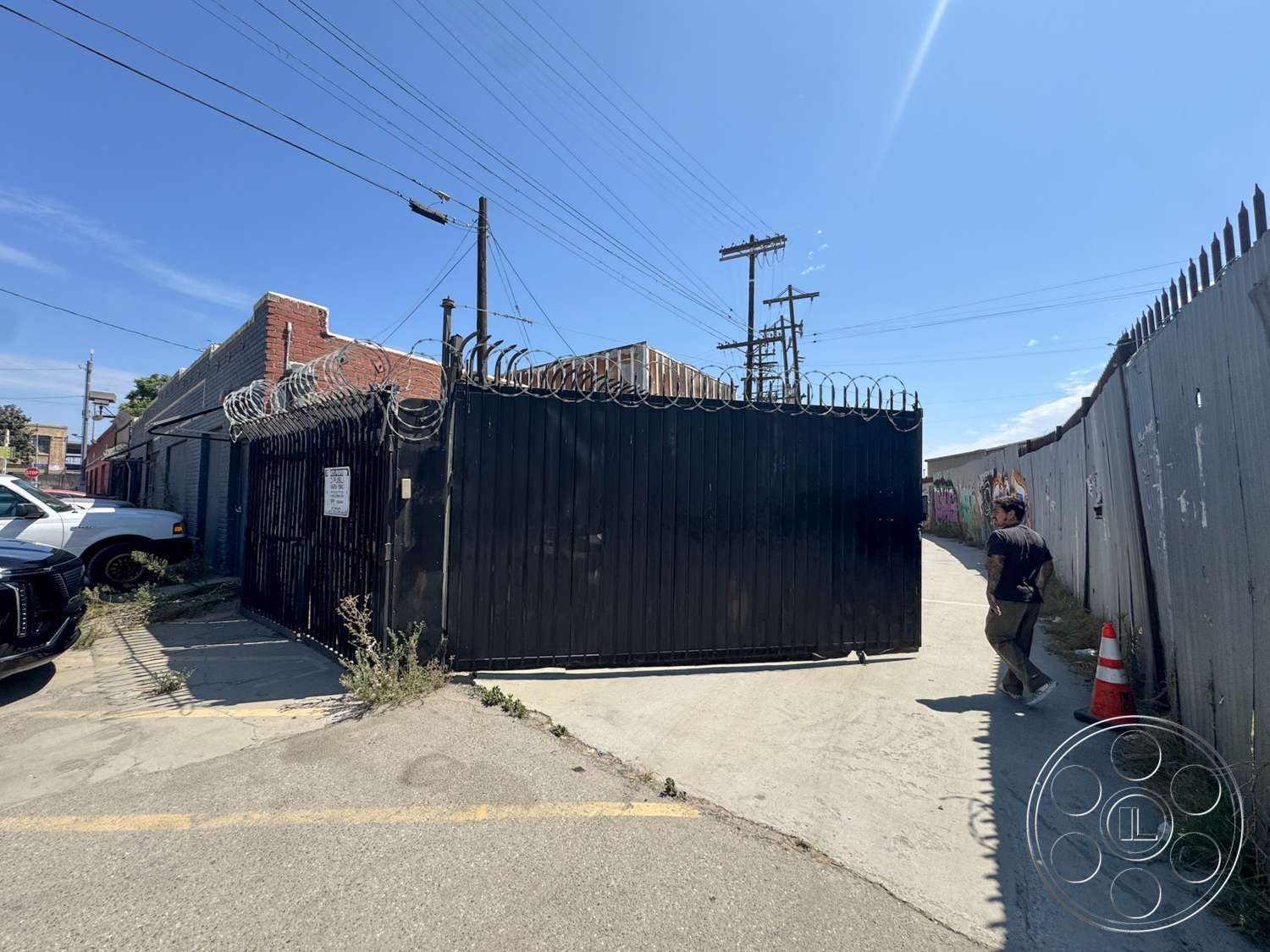 Warehouse 62 - exterior, black metal gate, chain link fence, industrial setting, brick building facade, overhead utility lines, gray concrete pavement, urban alleyway, signage on gate, barbed wire topping, painted graffiti wall, security cone, parked vehicle, sunlit sky
