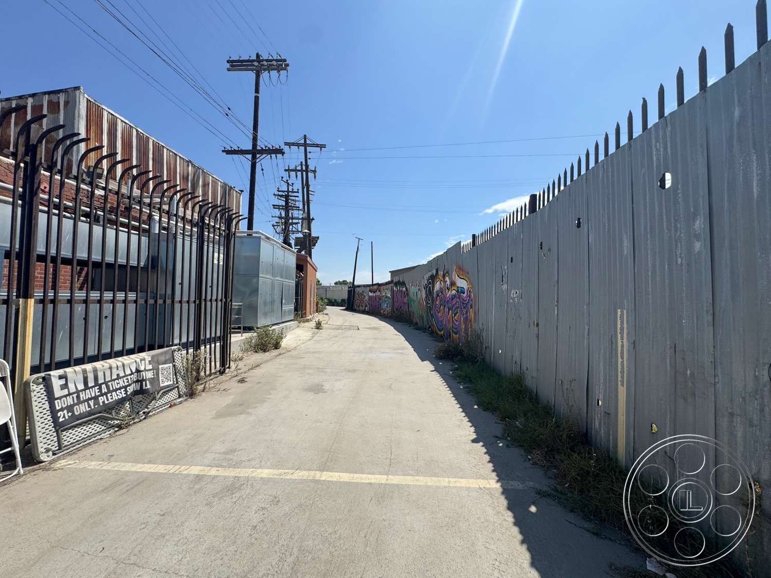 Warehouse 62 - exterior, dilapidated structures, chain link fence, graffiti wall art, bright blue sky, overhead power lines, urban industrial setting, brick building wall, weathered wooden fence, concrete alleyway pavement, metal corrugated fence, exposed red brick, scattered weeds and grass