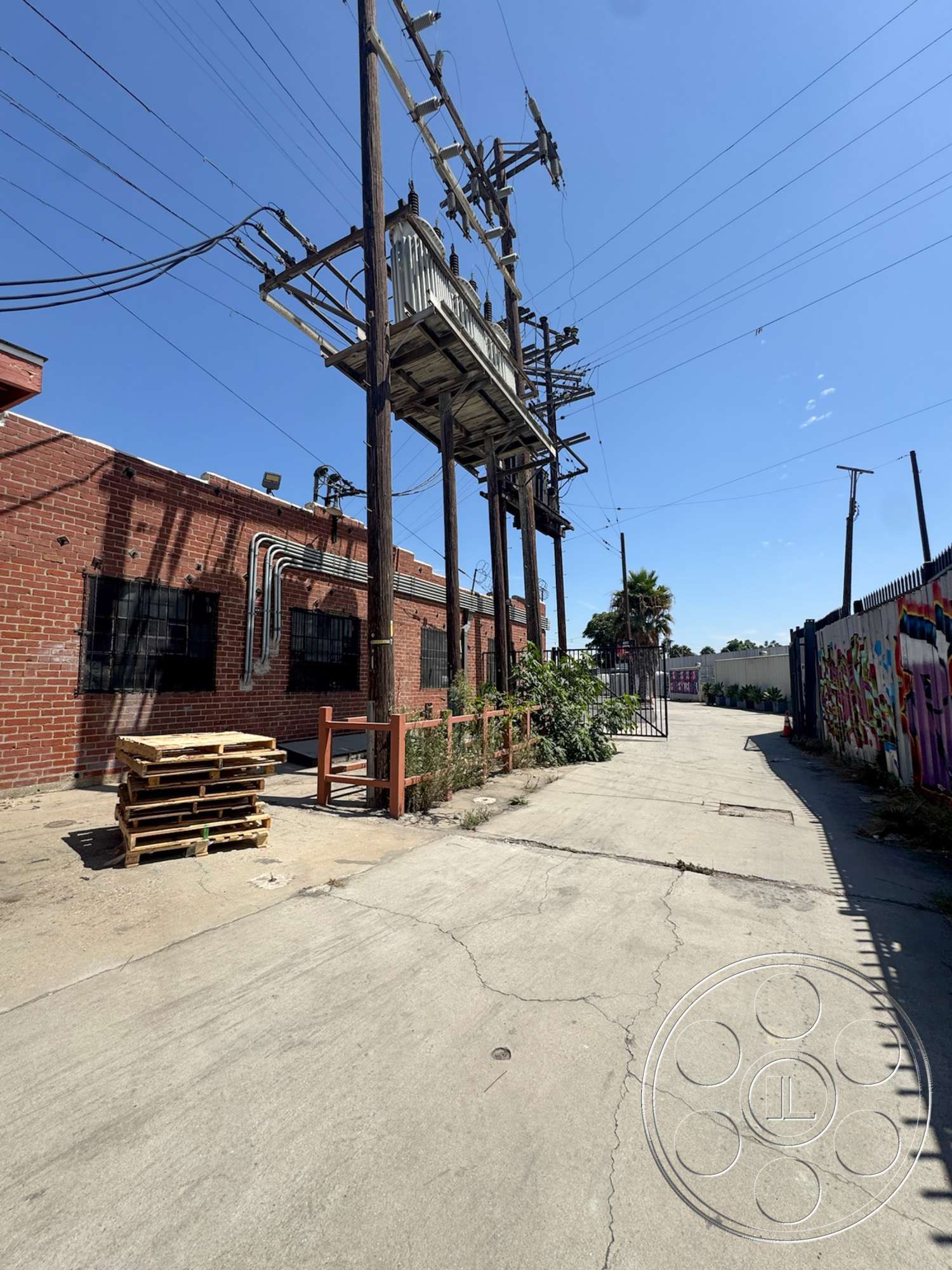 Warehouse 62 - outdoor greenery, overhead power lines, urban industrial setting, exposed brick warehouse walls, metal electrical poles, concrete alleyway, wooden pallet stacks, graffiti-covered exterior fence