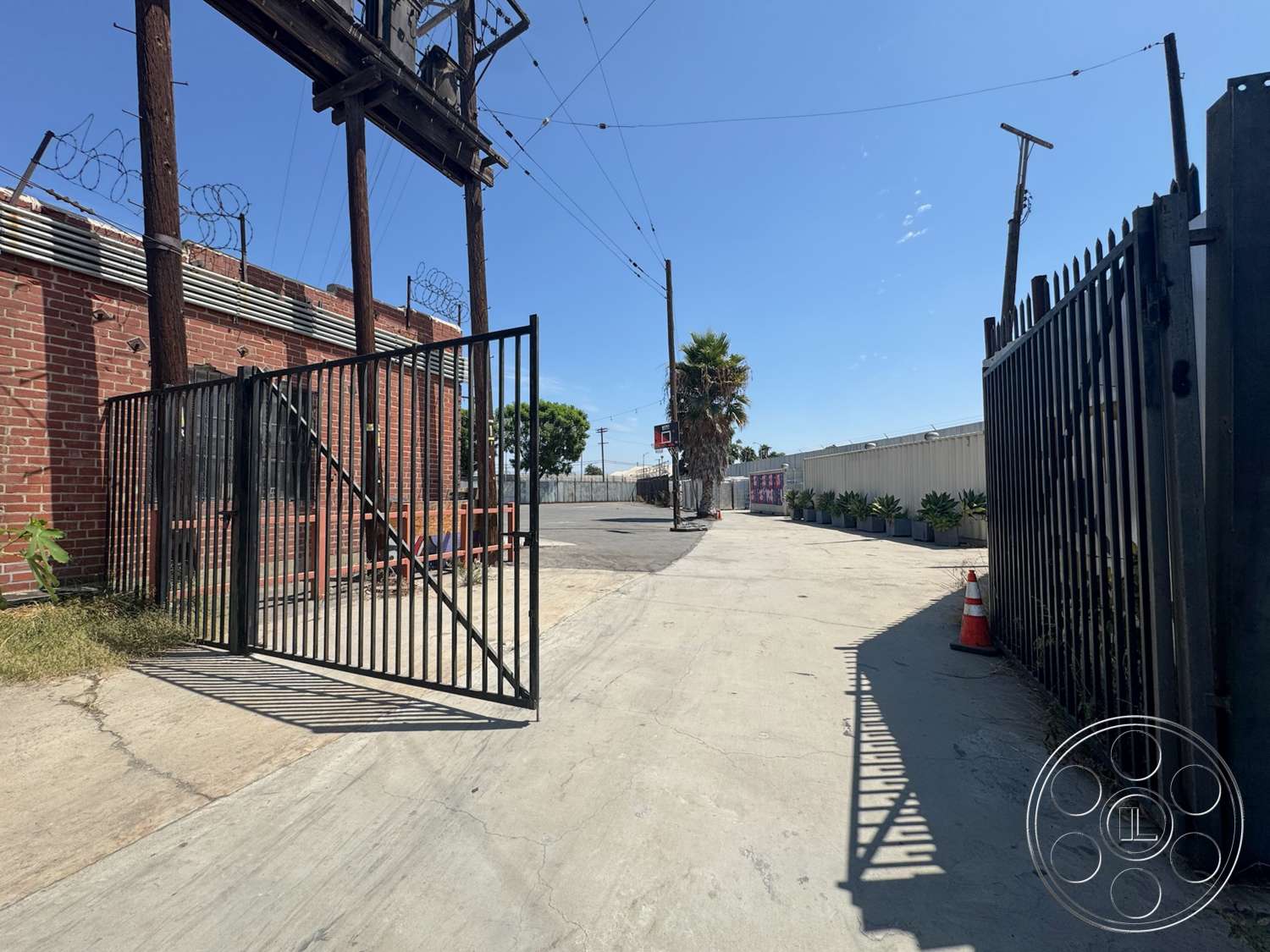 Warehouse 62 - exterior, potted plants, urban setting, brick wall, concrete driveway, palm tree, black metal gate, open sky, barbed wire fencing, paved roadway, pole-mounted signage