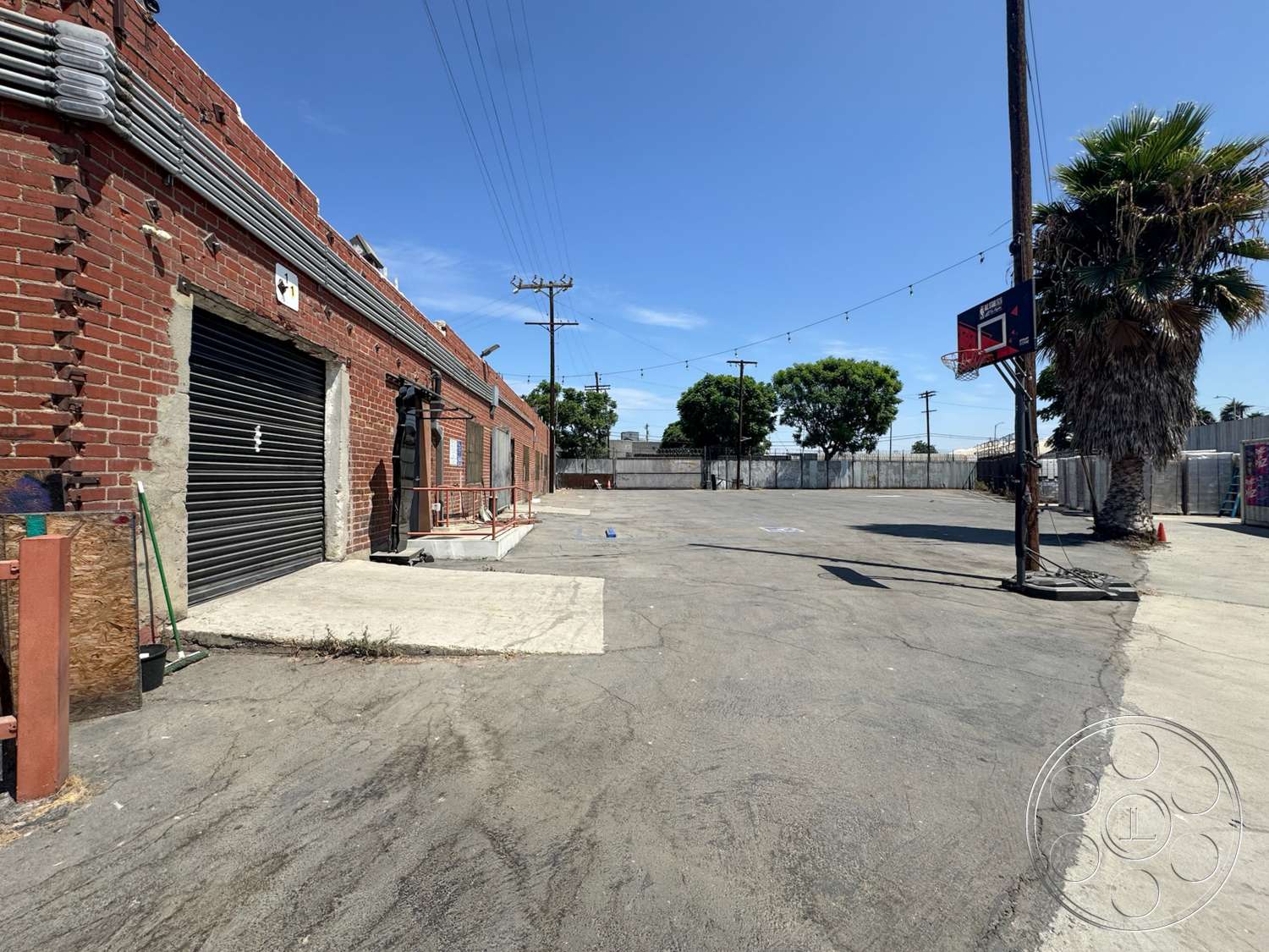 Warehouse 62 - exterior, concrete pavement, urban setting, industrial lighting fixtures, barrier fence, palm tree landscaping, overhead utility lines, metal roll-up garage door, exposed brick warehouse exterior, blacktop basketball court, telephone pole structure