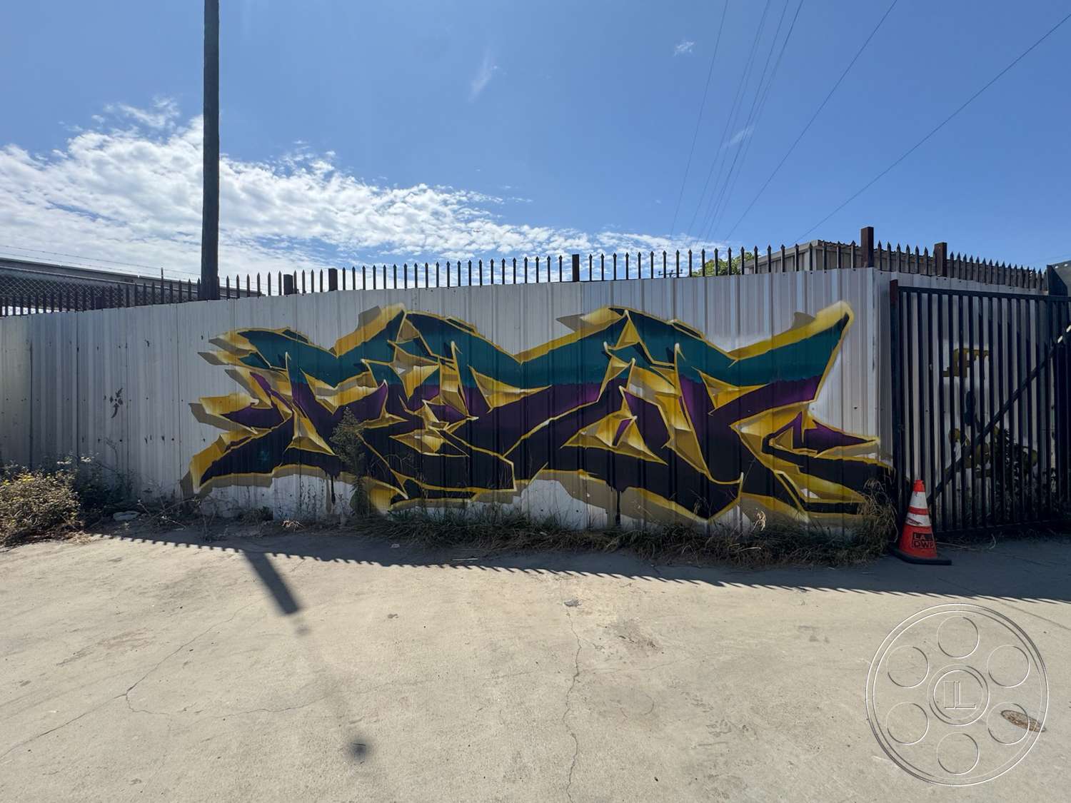 Warehouse 62 - exterior, concrete flooring, urban setting, metal fencing, blue sky, graffiti art, telephone pole, corrugated metal wall, overgrown grass, sunlit environment, industrial backdrop, colorful spray paint