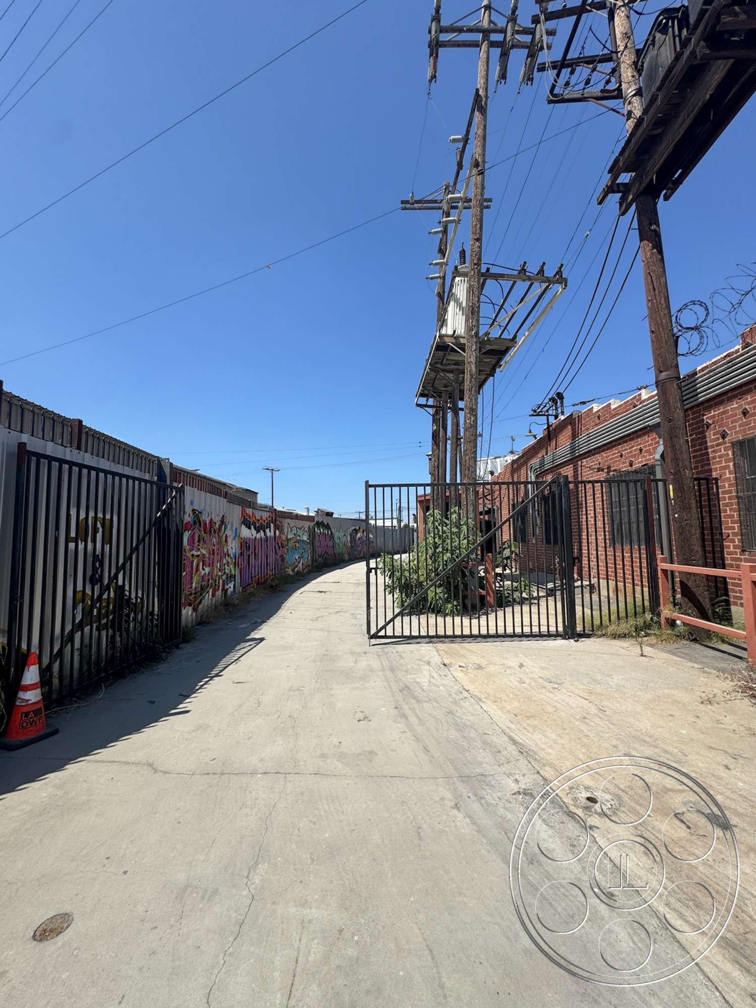 Warehouse 62 - exposed brick wall, metal gate, urban environment, asphalt pavement, bright blue sky, industrial setting, overhead power lines, graffiti-covered walls, barbed wire fencing, concrete alleyway