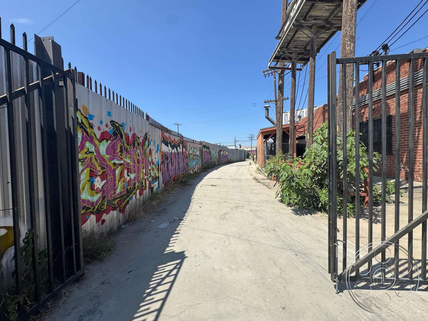 Warehouse 62 - exterior, concrete pathway, brick wall, metal security gate, urban alleyway, wooden utility poles, painted metal fence, graffiti art on fence, greenery along path