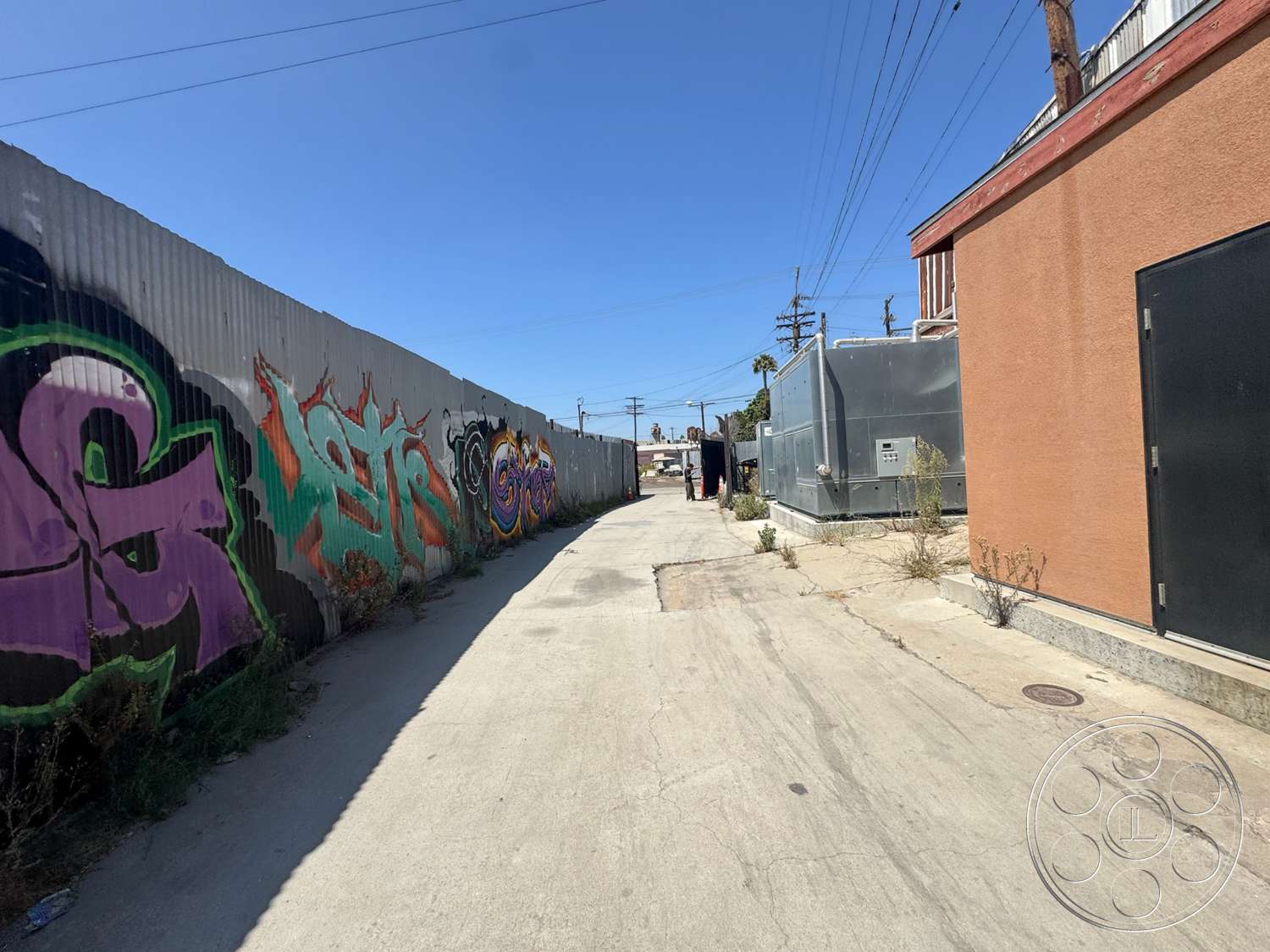 Warehouse 62 - exterior, dry vegetation, commercial buildings, utility equipment, industrial setting, flat concrete surface, blue sky backdrop, corrugated metal fence, concrete alleyway, colorful graffiti wall