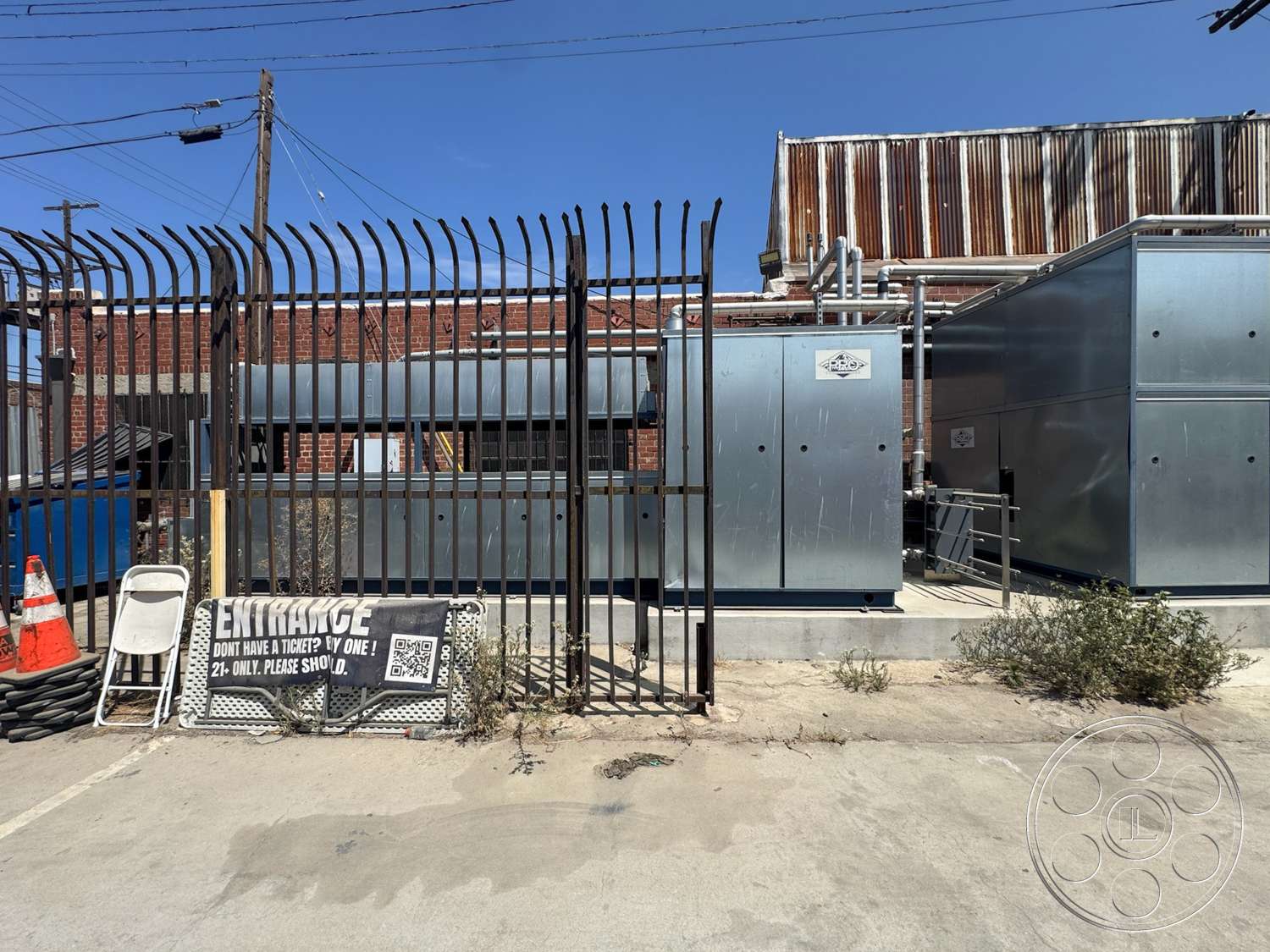 Warehouse 62 - exterior, concrete pavement, industrial setting, overhead utility lines, metal fence with vertical slats, signage with bold typography, steel utility boxes, brick wall with exposed masonry, weathered metal roof, plant growth along pavement