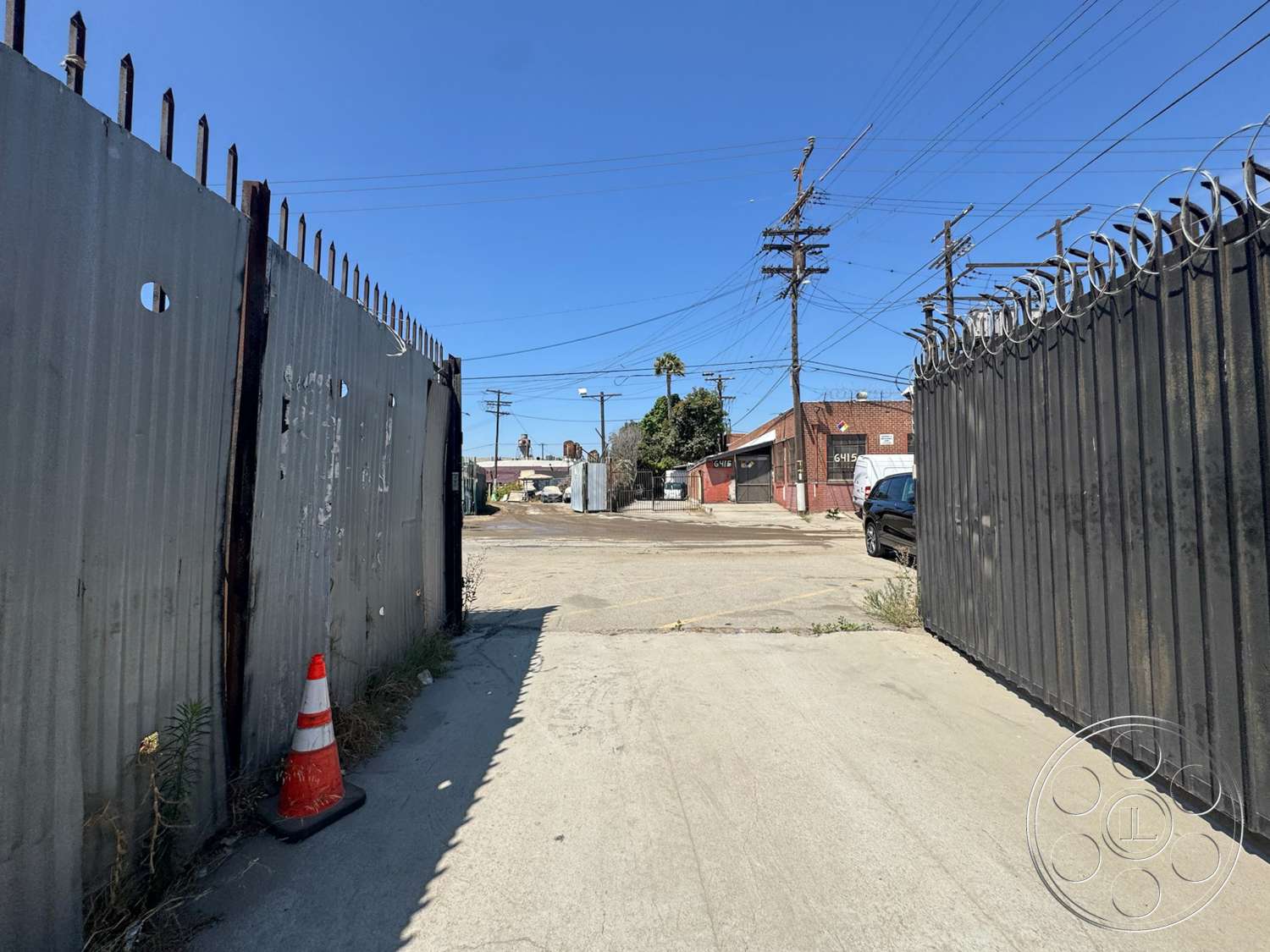 Warehouse 62 - exterior, urban landscape, concrete driveway, utility poles, pavement markings, traffic cone, industrial setting, overhead power lines, barbed wire details, gravel ground surface, rusted metal fencing, corrugated steel gate