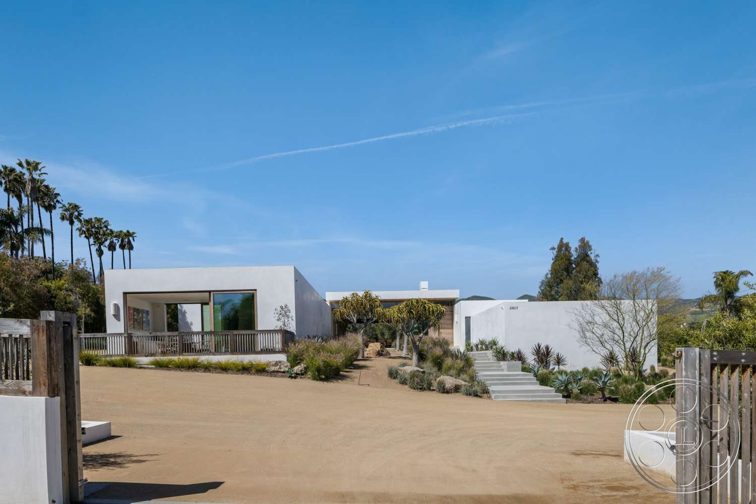 Modern 529 - exterior, natural stone accents, white stucco walls, modern architectural style, large floor-to-ceiling windows, dirt driveway, desert setting, flat roof design, landscaped garden area, wooden deck railing, concrete steps leading to entrance, palm trees in surroundings
